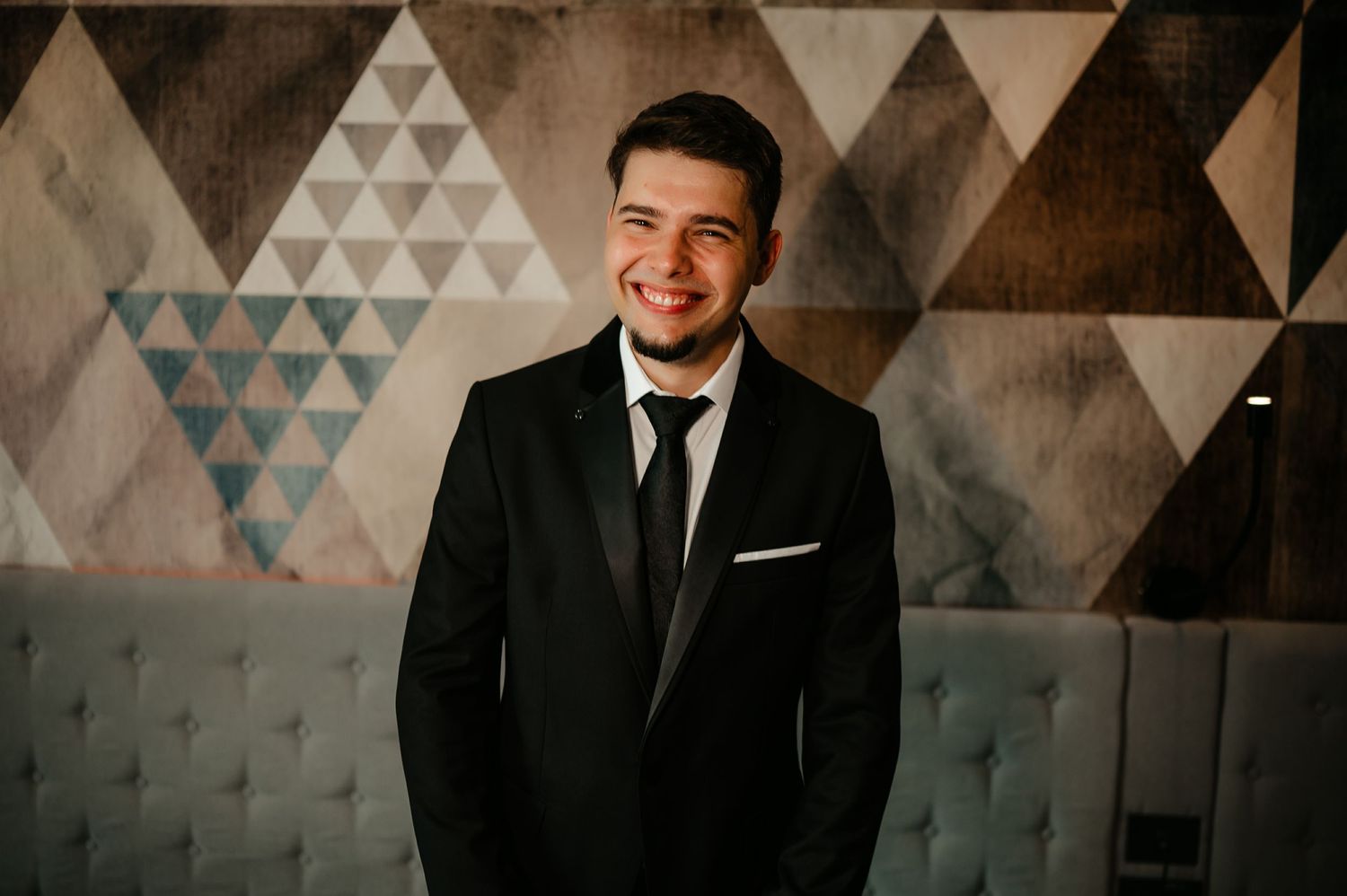 Smiling young man in black tuxedo suit with tie standing before geometric patterned wall.