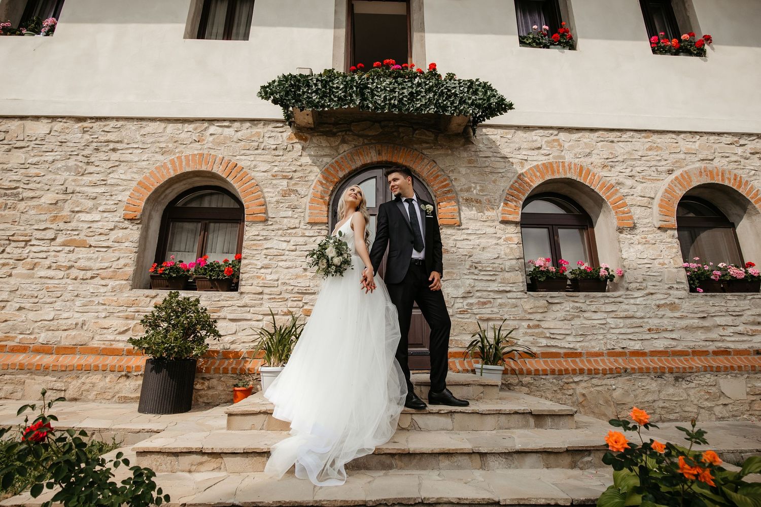 Bride and groom pose on stone steps outside a rustic venue adorned with flower boxes and arched windows.