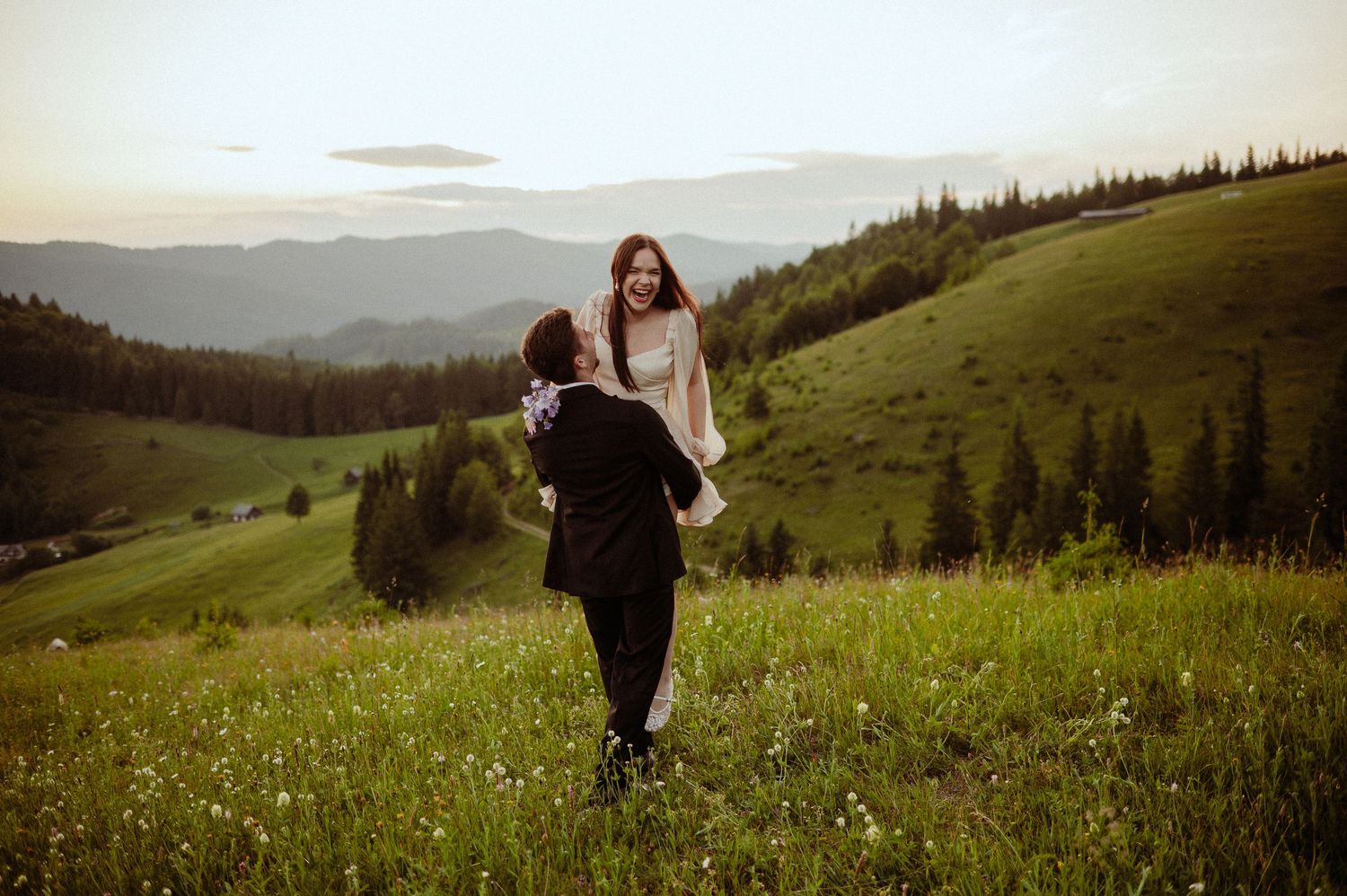Couple sharing a romantic moment on a lush green hillside at sunset with mountain views.