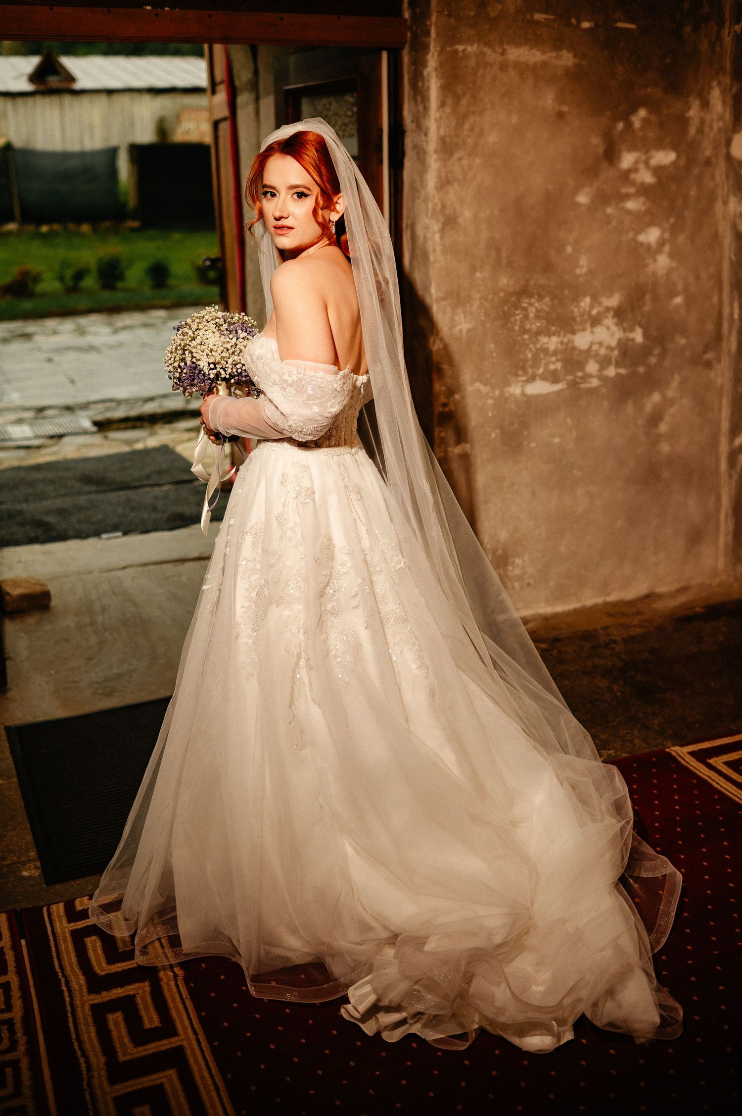 Redhead bride in off-shoulder lace wedding gown with veil holds bouquet while posing in doorway.