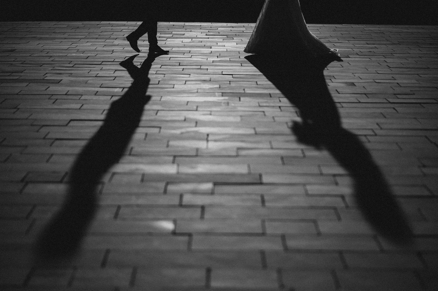 Dramatic black and white silhouette of bride and groom walking on cobblestone with long shadows at night.
