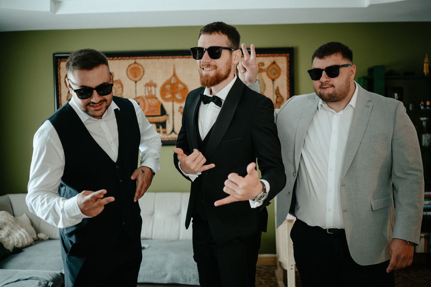 Three groomsmen in formal attire posing with sunglasses and fun gestures in a hotel room.