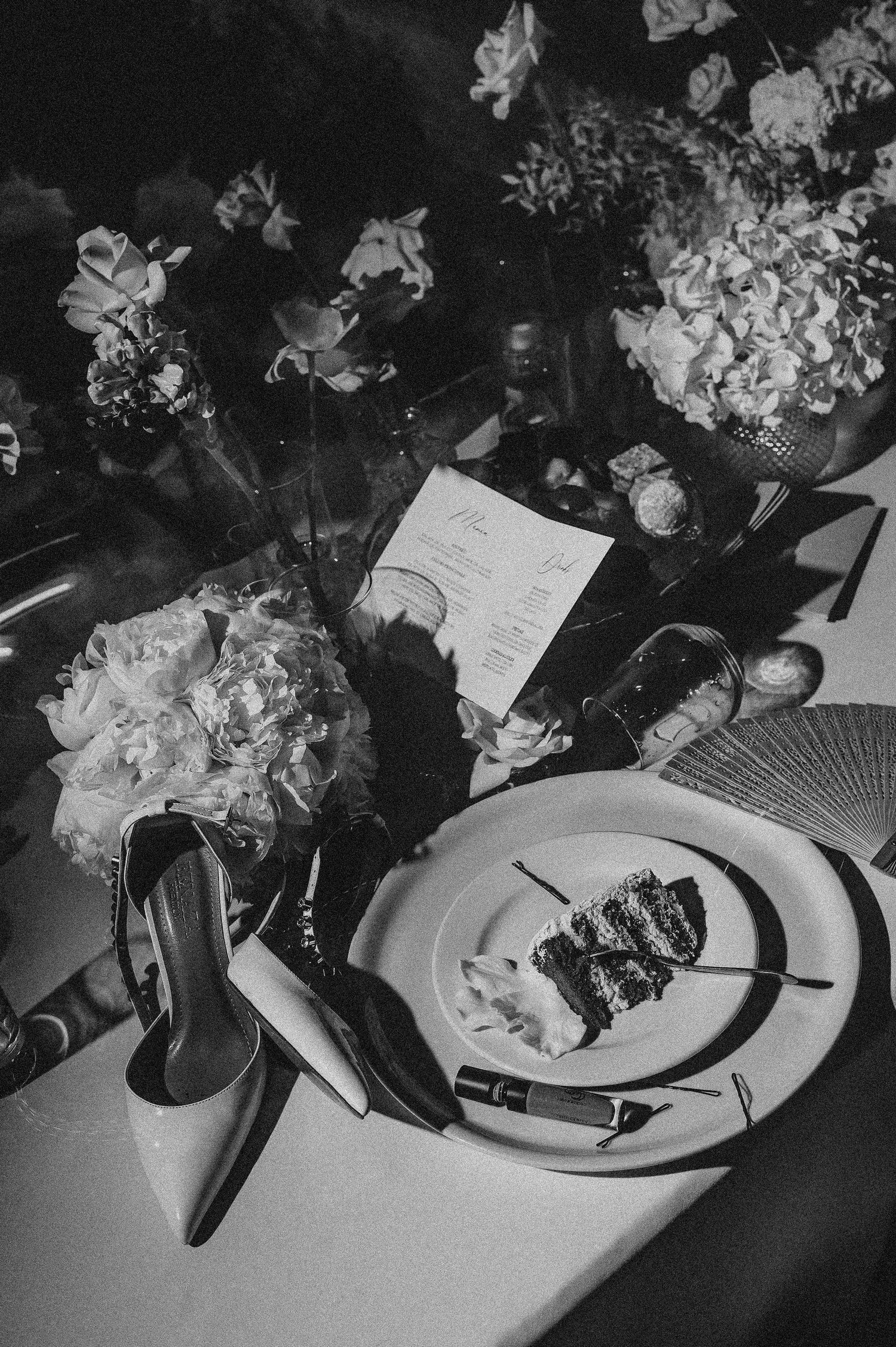 Black and white flat lay of wedding table with cake slice, high heel shoes, flowers, and menu card.