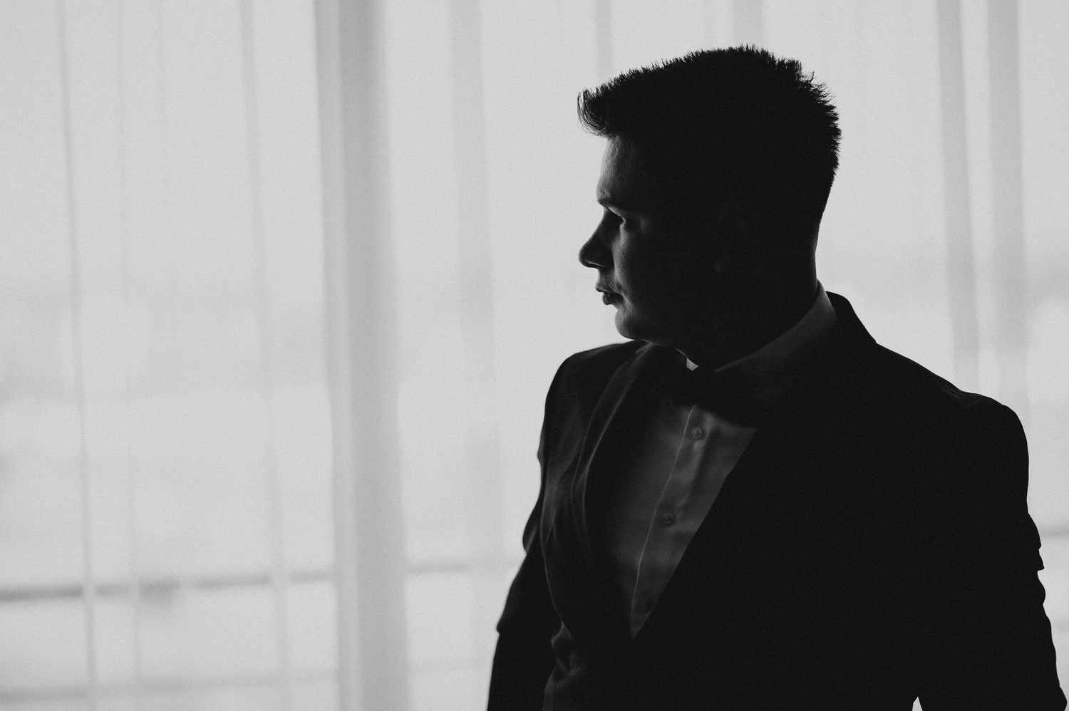 Dramatic black and white silhouette portrait of groom in tuxedo against bright window curtains.