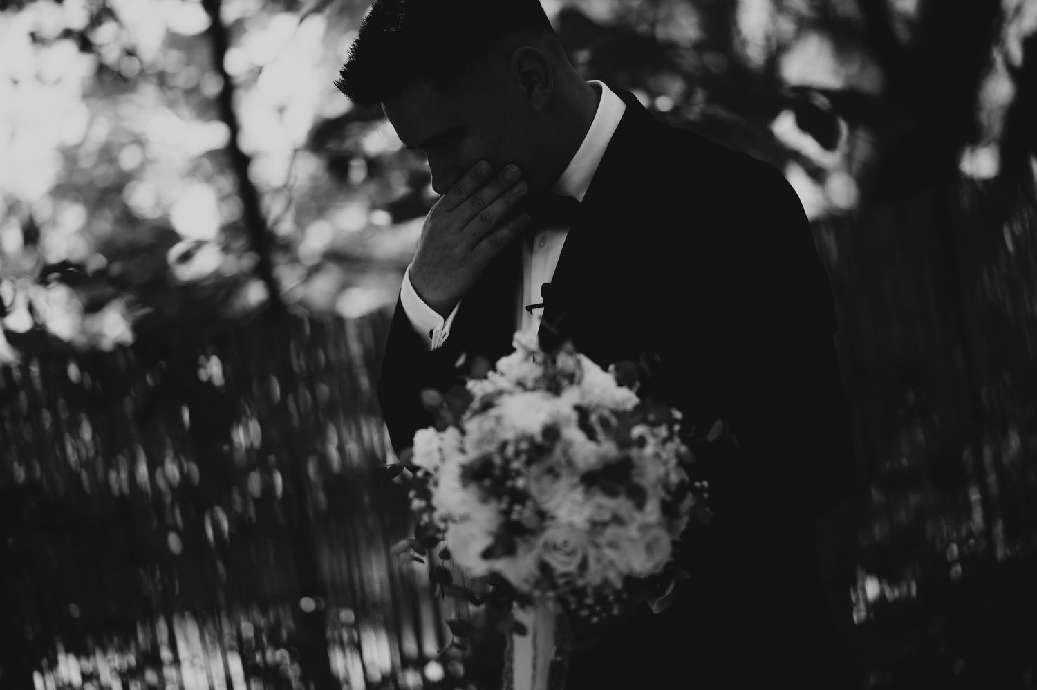 Emotional groom in black tuxedo holding bouquet, black and white wedding photography.