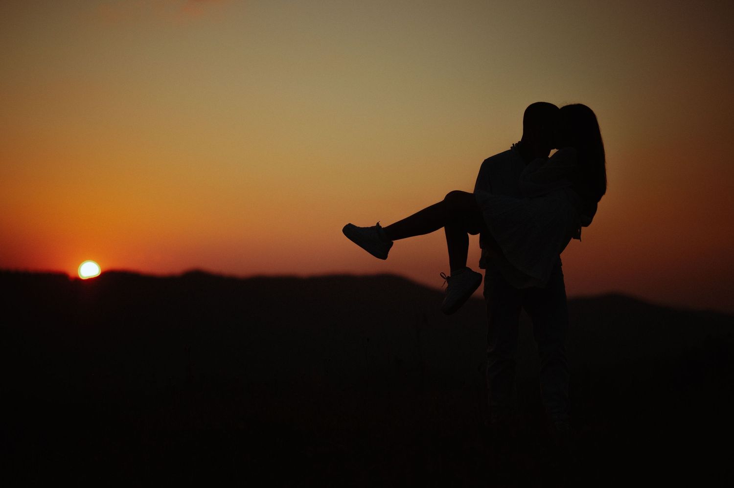 Romantic silhouette of man lifting woman against a vivid orange sunset over mountains.