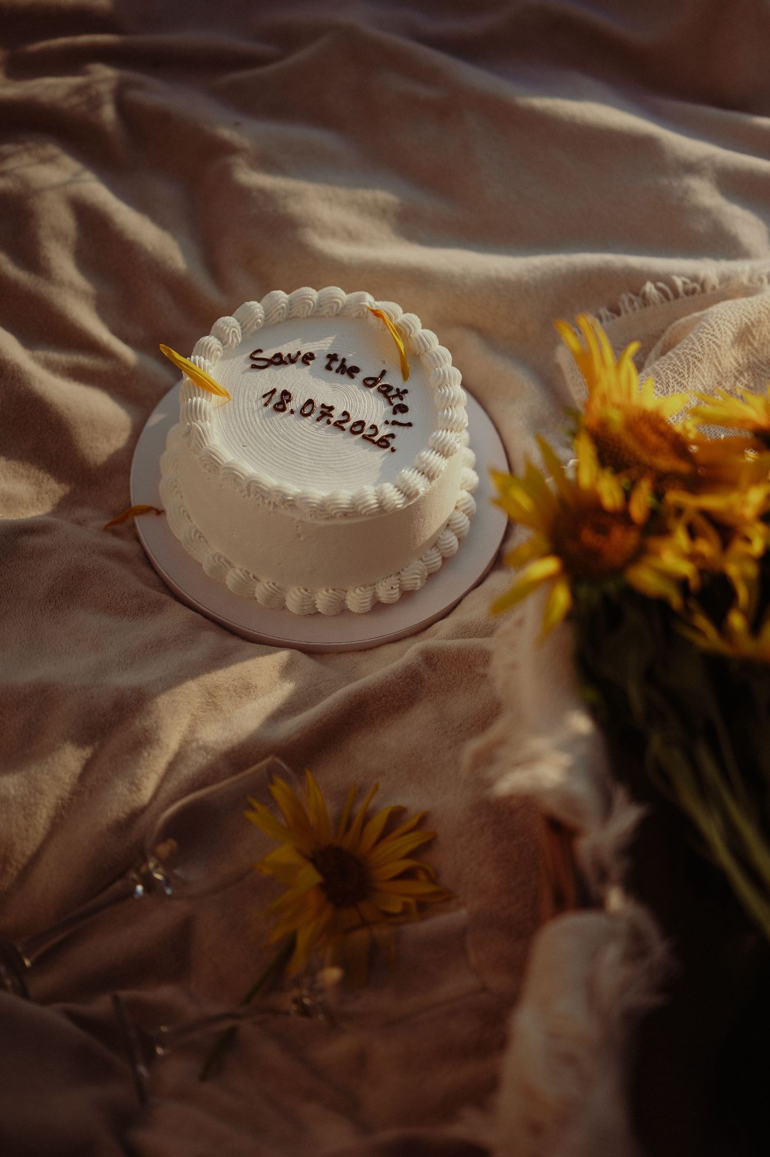 White round cake with Save the Date inscription surrounded by sunflowers on cream fabric.