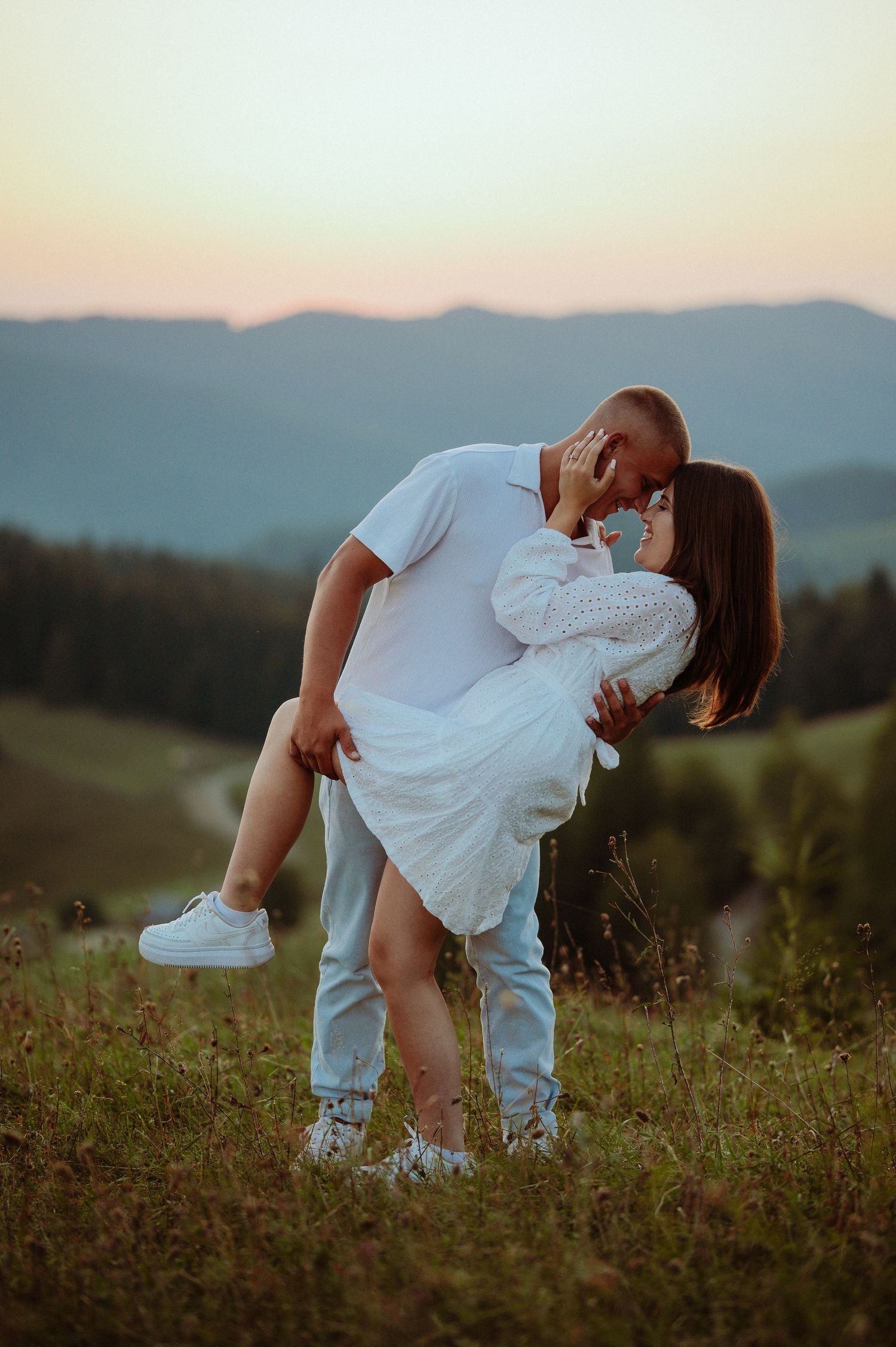Couple sharing a romantic dip kiss on green hillside at sunset with mountain landscape in background.
