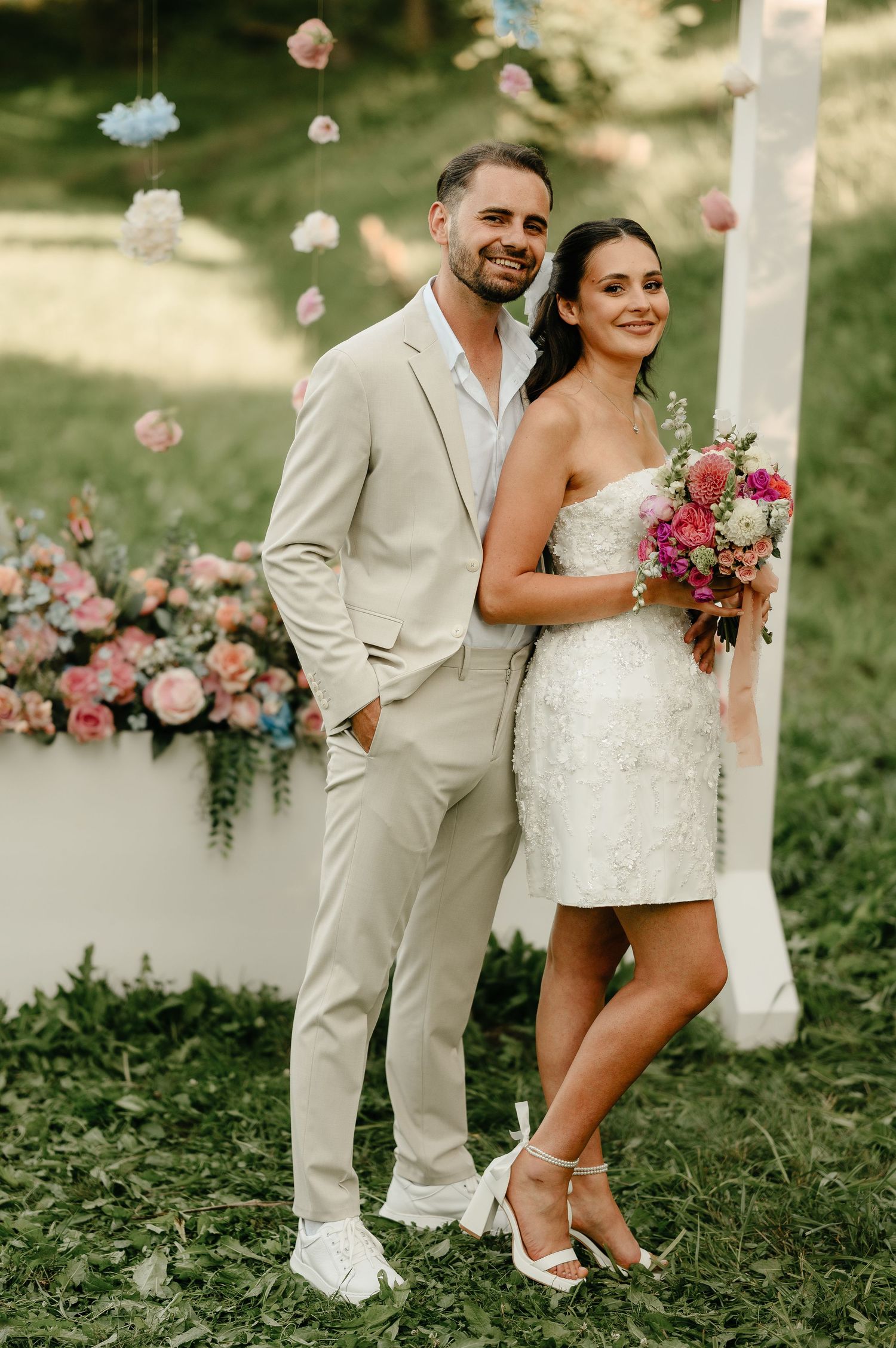 Bride and groom pose together at outdoor wedding with pink floral arch and hanging decorations.