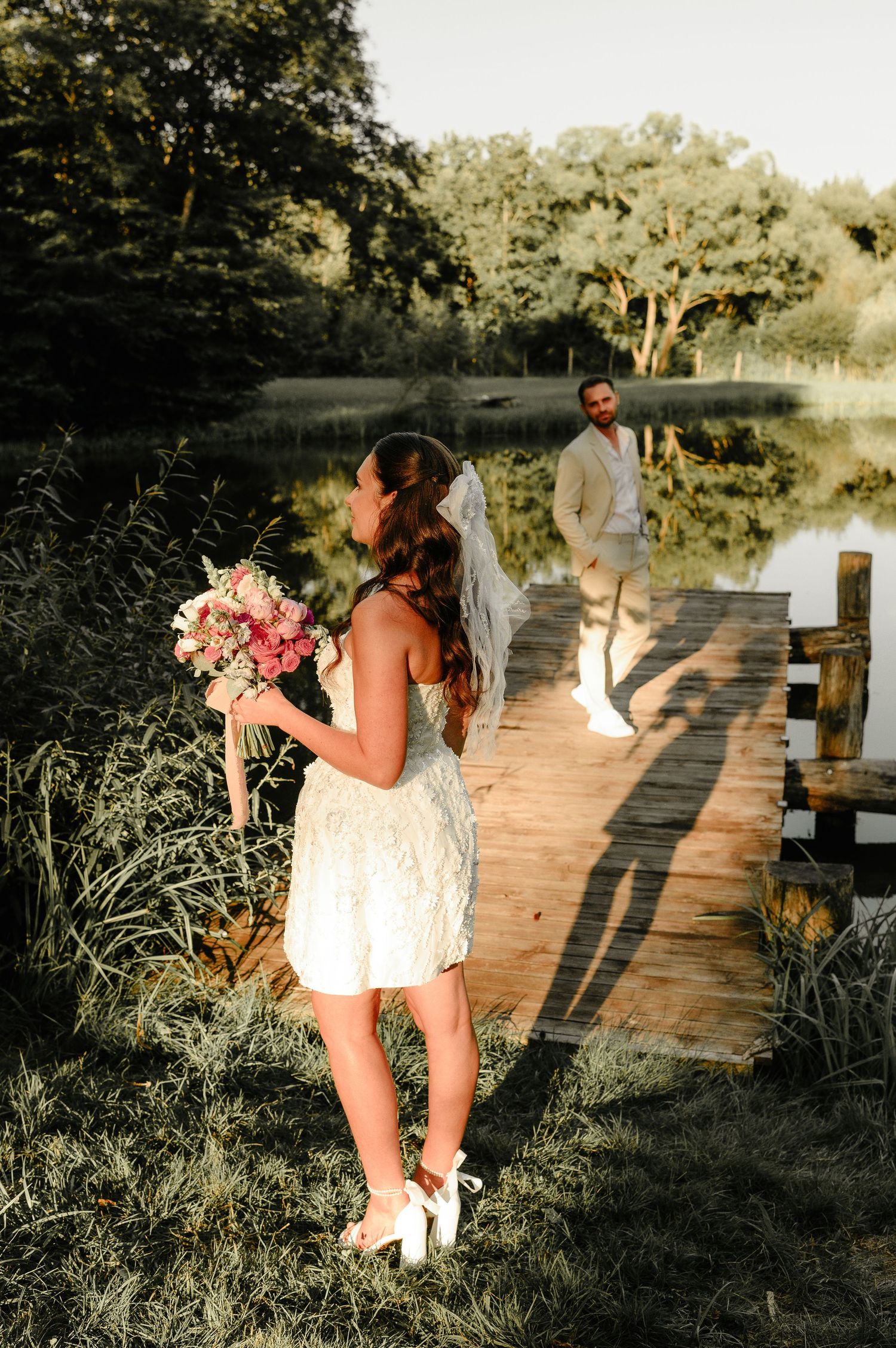 Bride with veil bow holds pink bouquet near lakeside dock as groom stands in background at sunset.