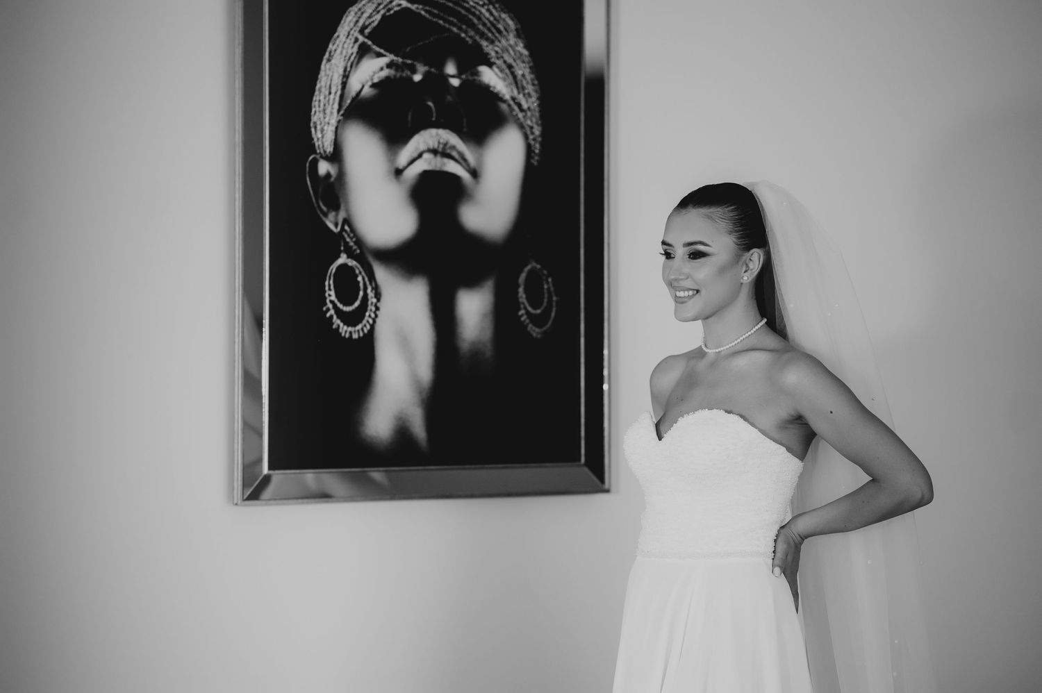 Bride in strapless white gown smiling beside framed artwork, black and white.