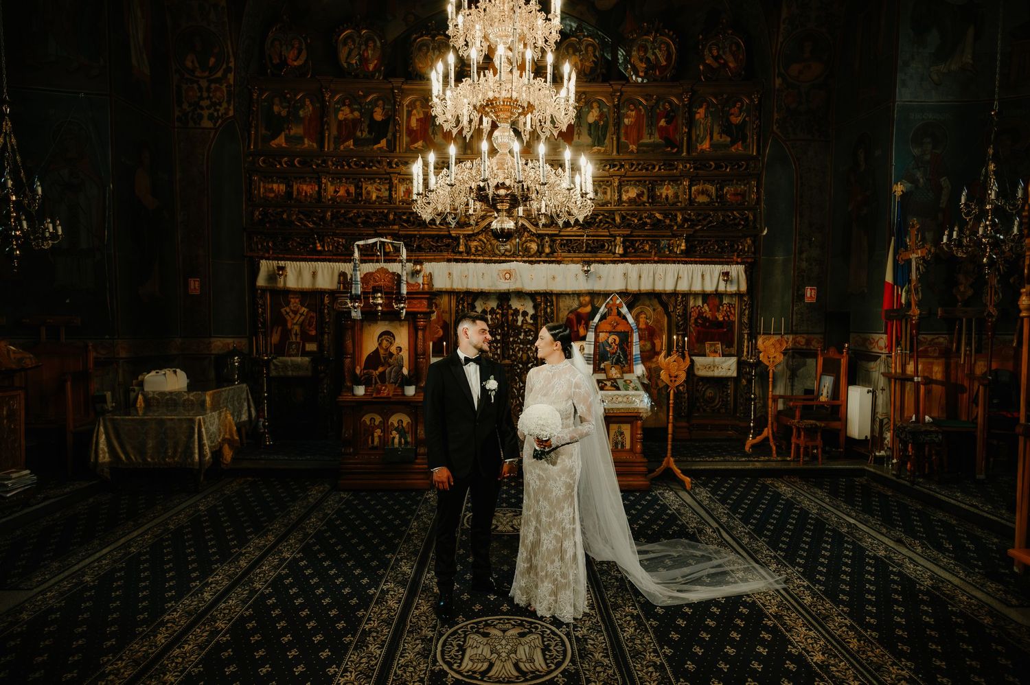 Bride and groom standing together beneath a chandelier in a beautifully decorated Orthodox church.