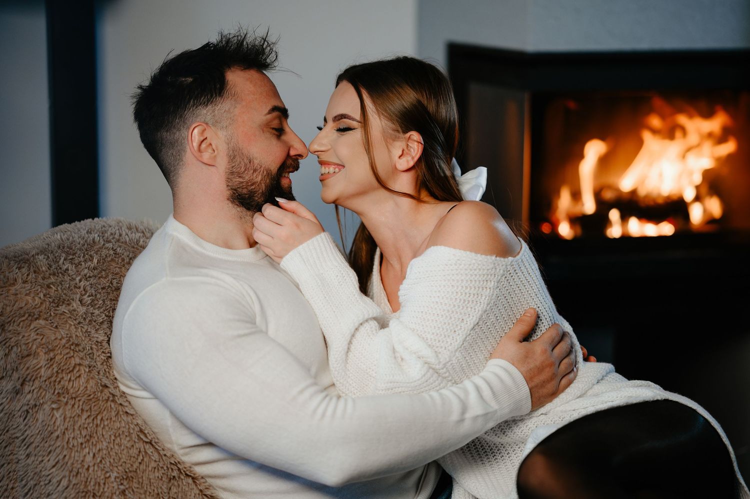 Loving couple in white sweaters cuddle and smile by a glowing fireplace indoors.