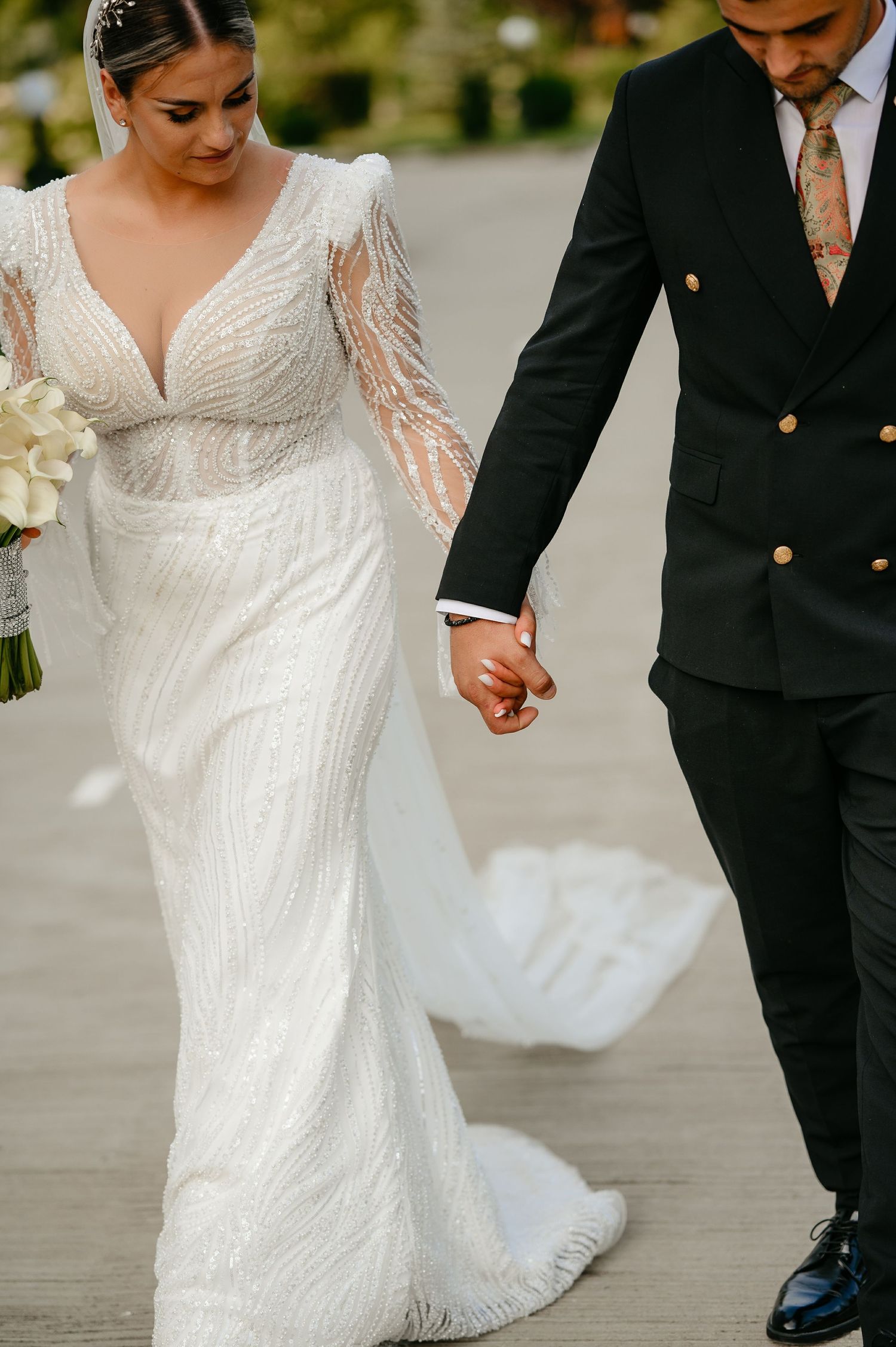 Bride in long-sleeve beaded wedding gown with train holds hands with groom in black suit while walking outdoors.