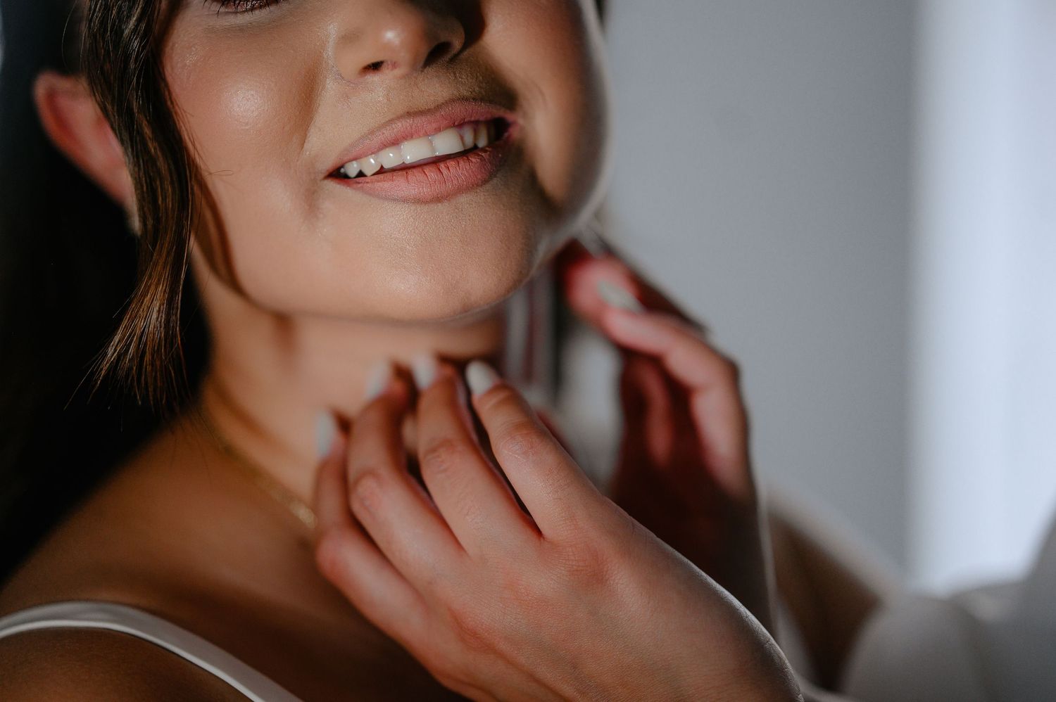 A smiling bride having a delicate necklace fastened around her neck during wedding preparations.