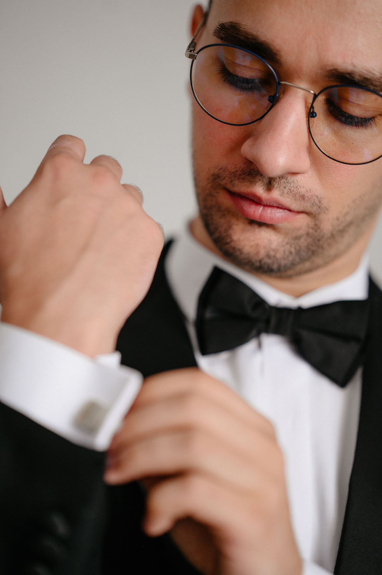 Close-up of groom in black tuxedo fastening cufflinks, wearing round glasses, black and white portrait.