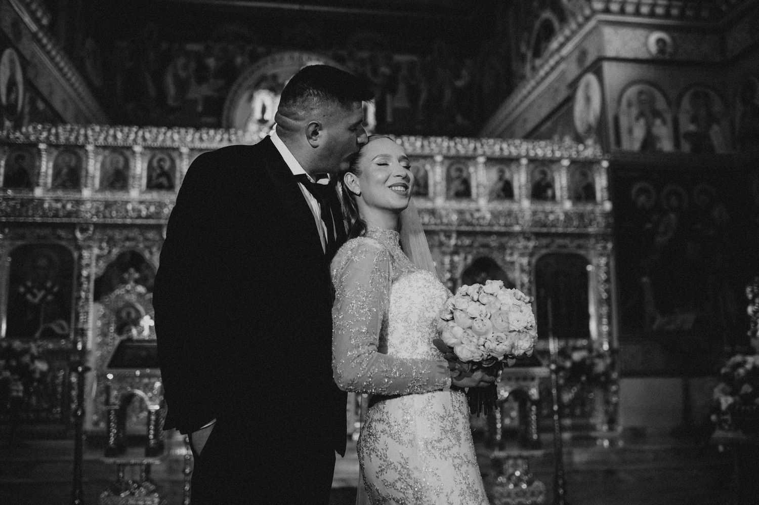 Bride and groom share romantic moment in ornate Orthodox church in black and white photo.