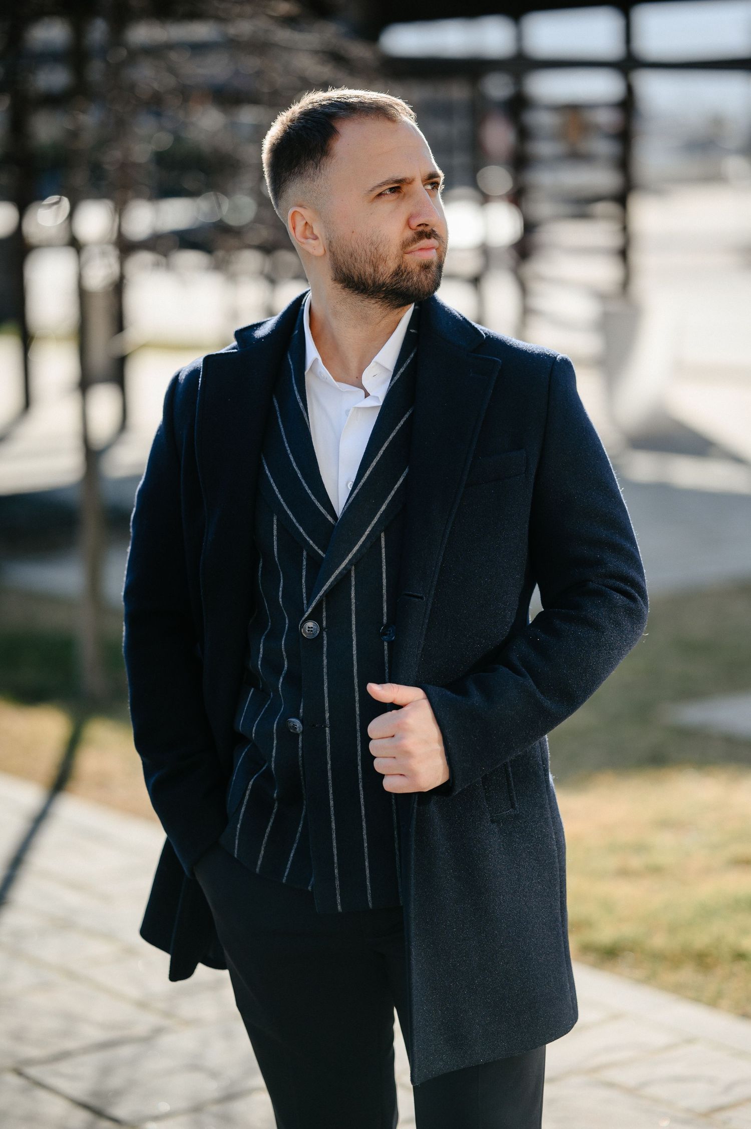 Dapper bearded groom in dark navy overcoat and striped suit poses outdoors in sunlight.