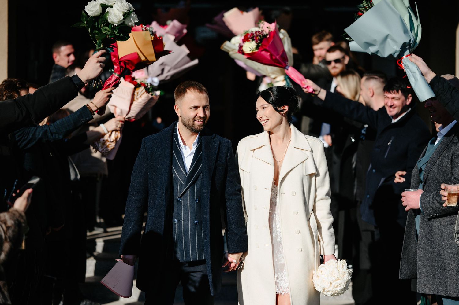 Newlywed couple walks through a celebratory arch of flowers held by guests outside a wedding venue.