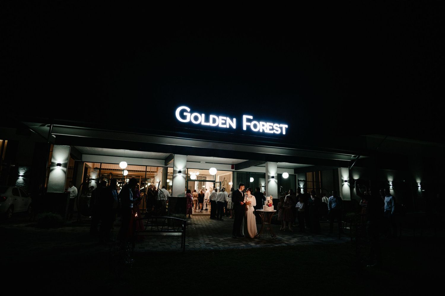 Nighttime exterior of Golden Forest venue with illuminated sign and guests gathered outside.