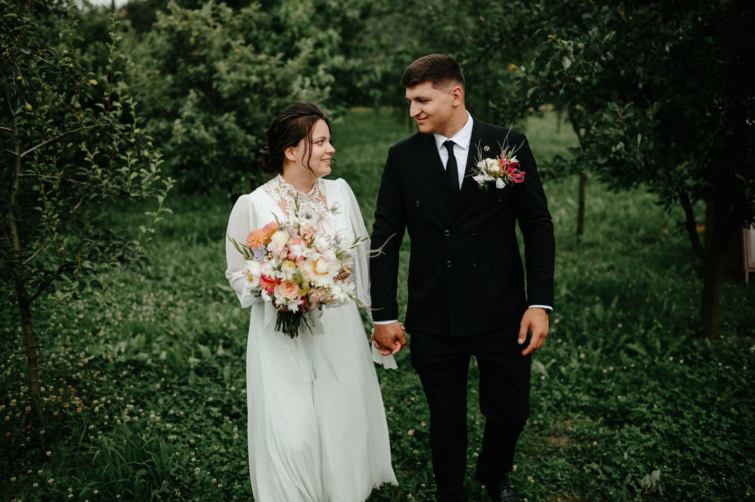 Bride and groom walk hand in hand through lush green garden, bride holding colorful bouquet in white gown.