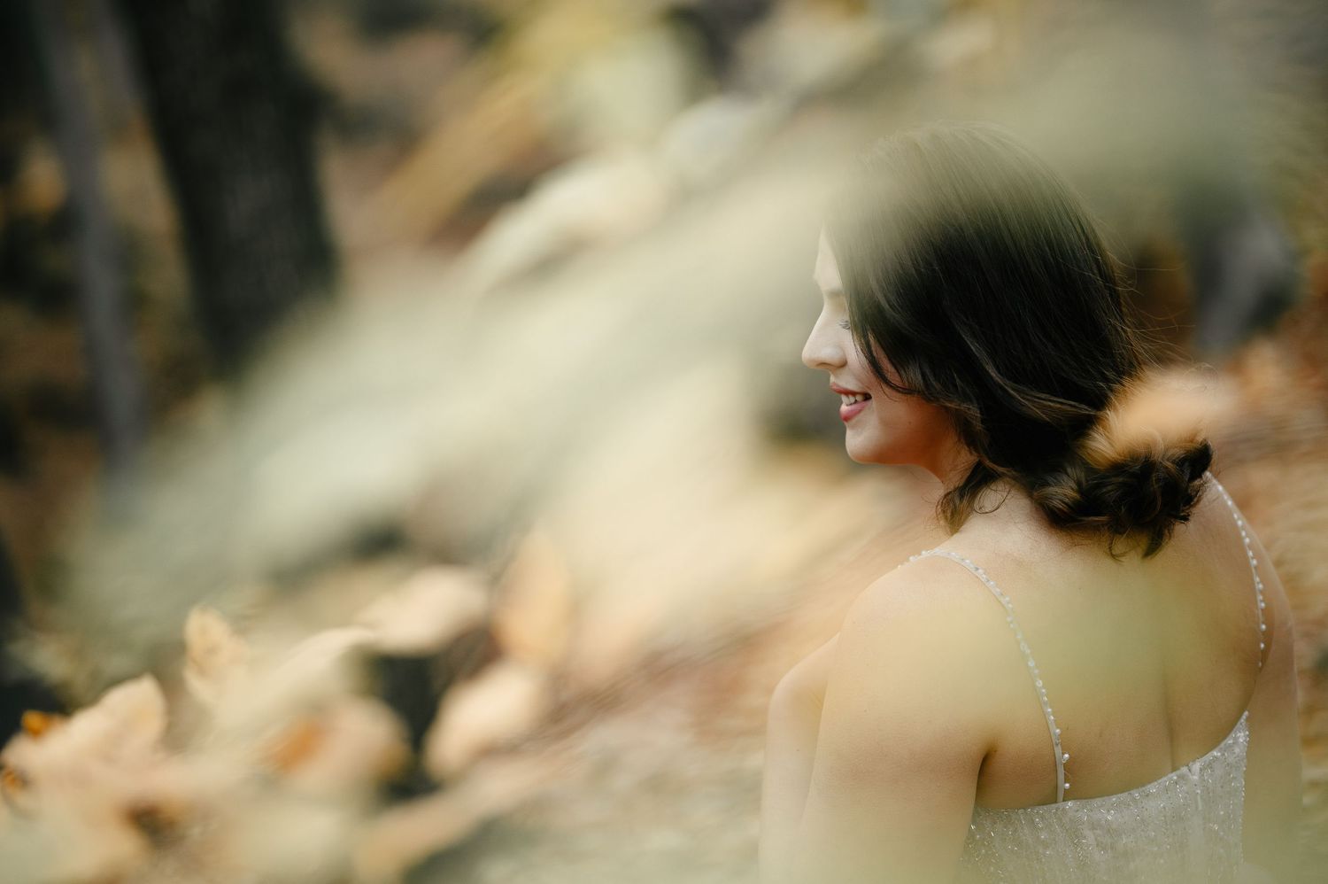 A woman in a white dress smiles softly in an autumn forest, captured through blurred golden leaves.
