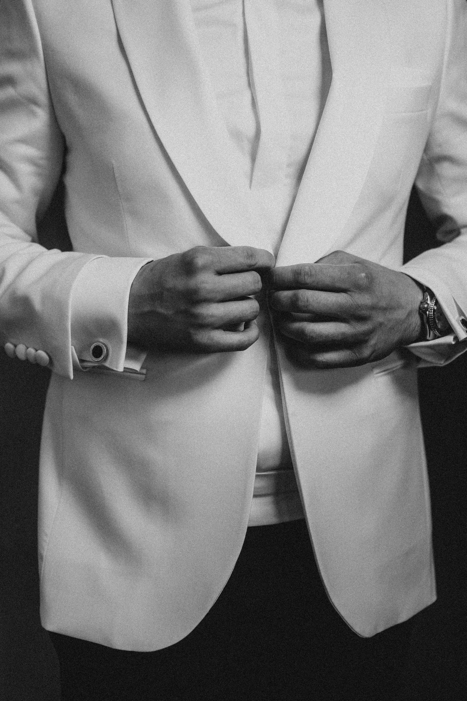 Black and white photo of a groom buttoning his white tuxedo jacket, showcasing elegant cufflinks and a watch.