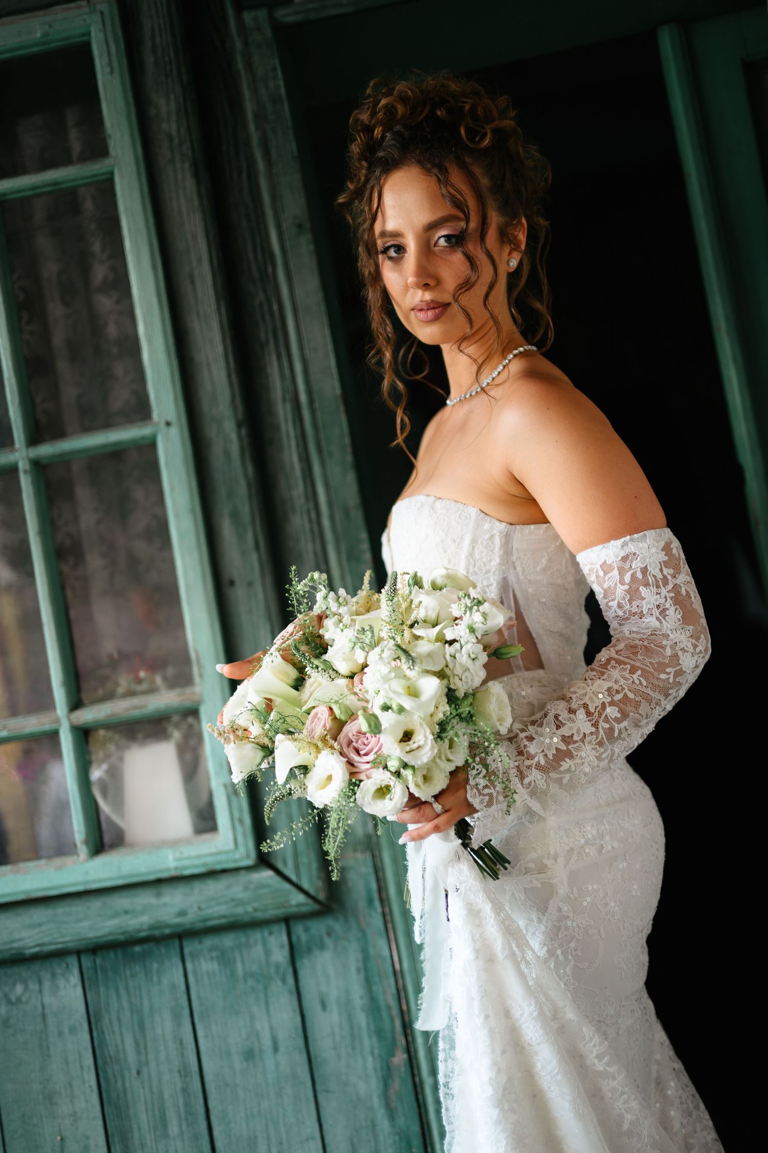 Elegant bride in lace wedding dress and gloves holding white floral bouquet standing by rustic teal wooden door.