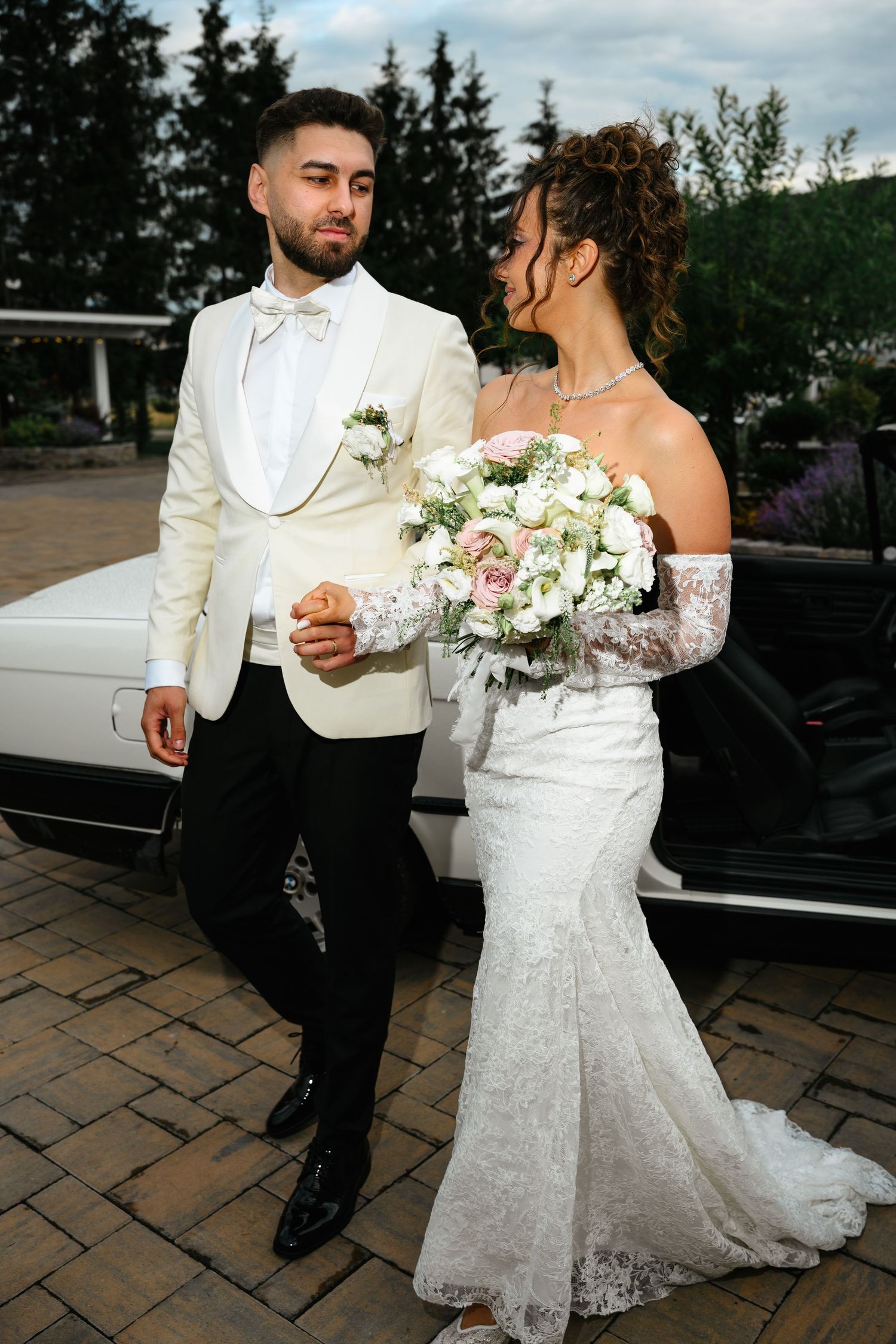 Elegant bride in lace gown holding floral bouquet stands with groom in white tuxedo jacket by a white car.