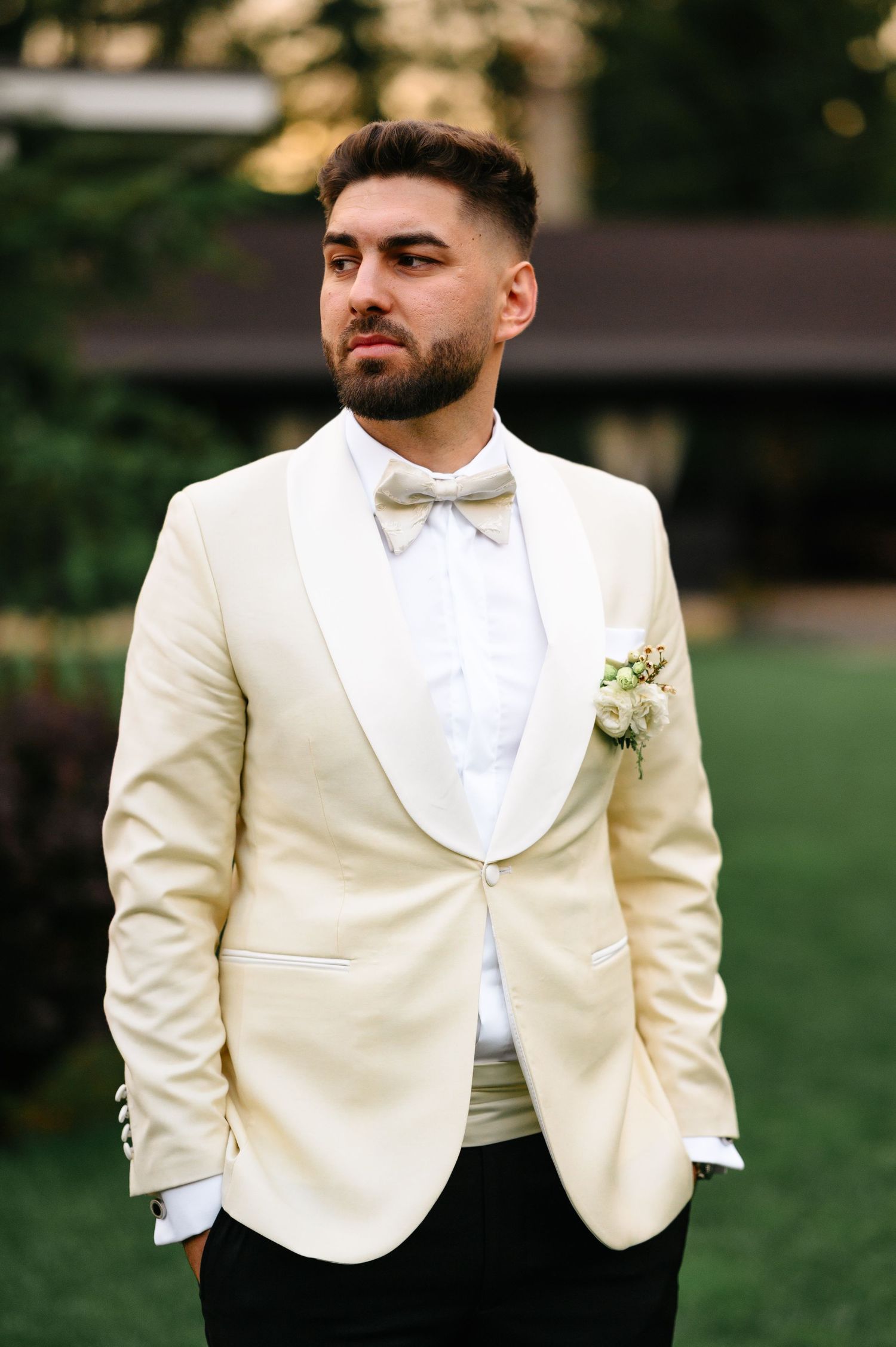 Groom in cream tuxedo jacket with bow tie and boutonniere poses outdoors at wedding venue.