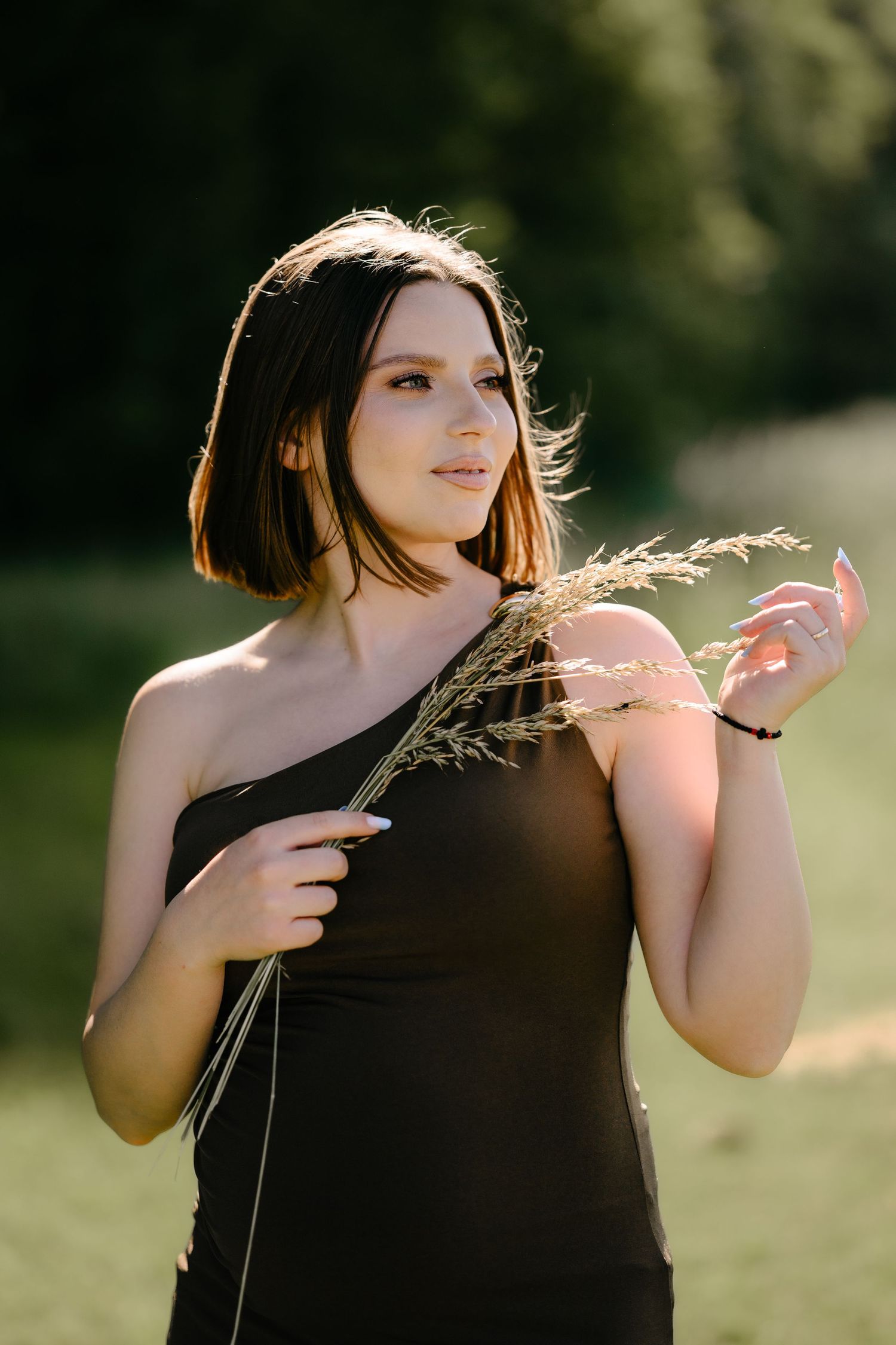 Pregnant woman in dark one-shoulder dress holding dried grass in golden sunlight outdoors.