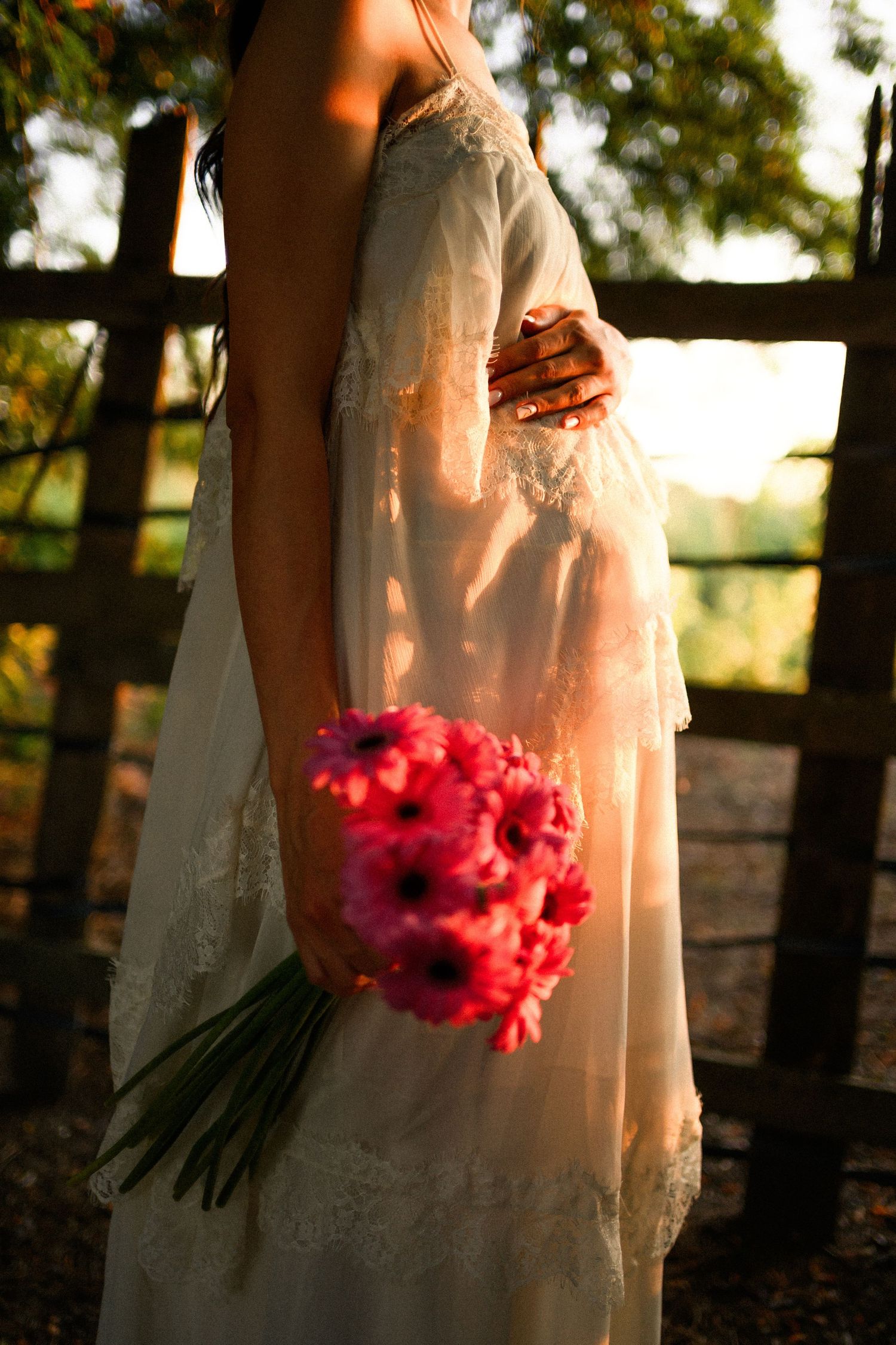 Pregnant woman in white lace dress holding pink gerbera bouquet at golden hour near wooden fence.