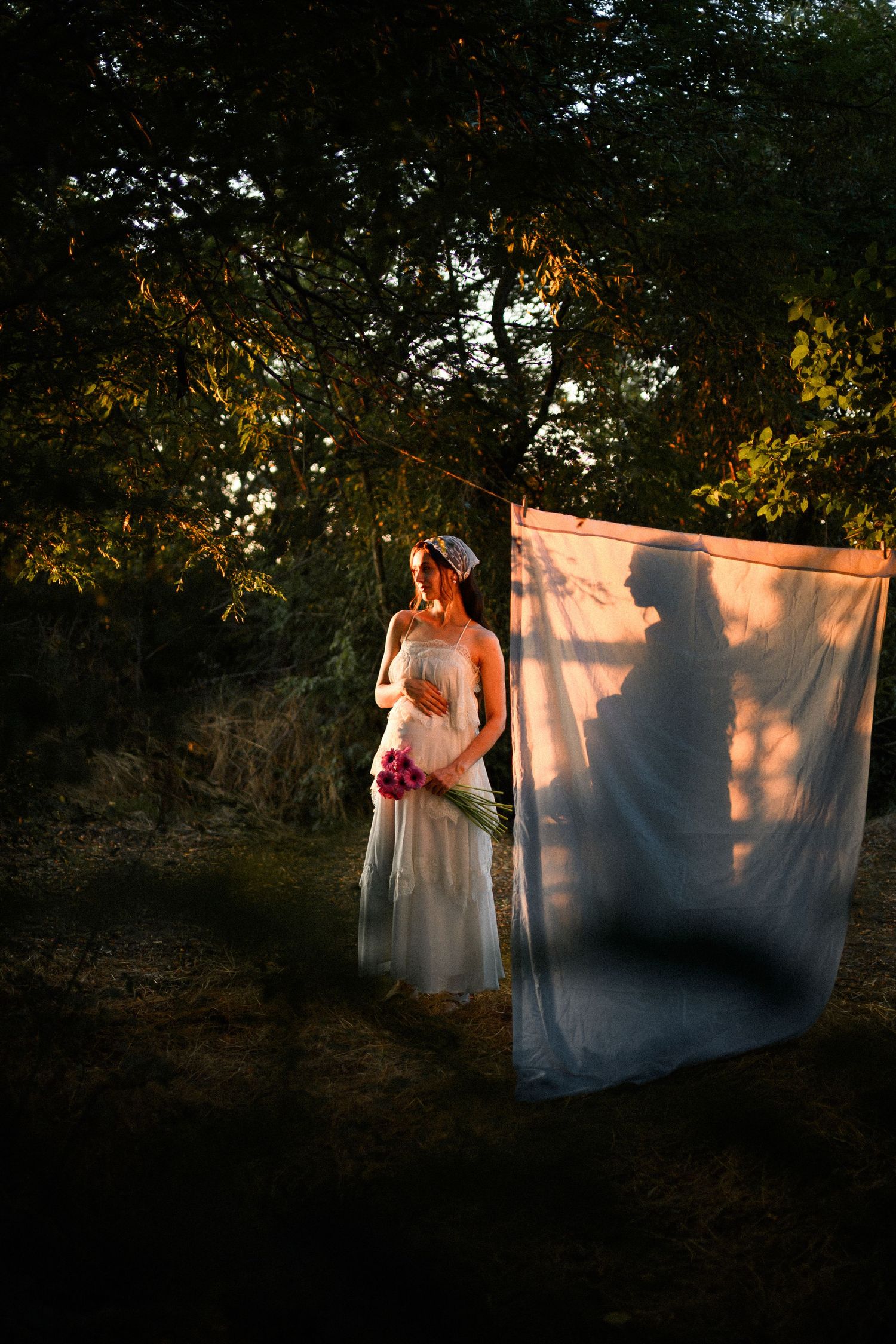 Bride in white gown holding bouquet stands by sheer fabric panel in forest, with shadowy figure projected on cloth.