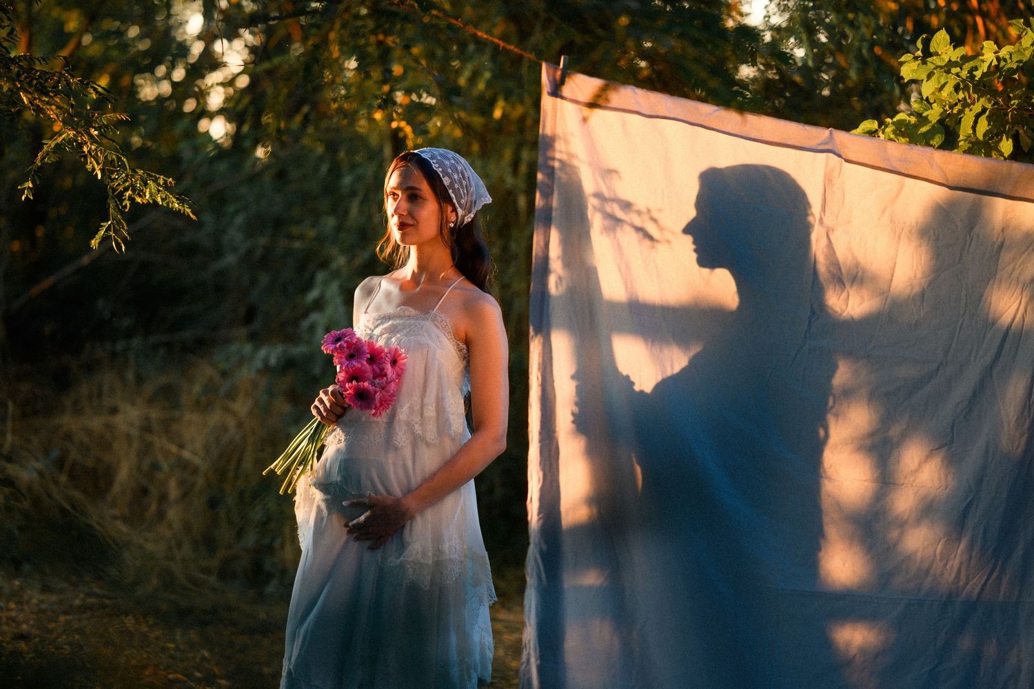 Pregnant woman in white dress holding pink flowers, with her shadow cast on a white sheet outdoors.