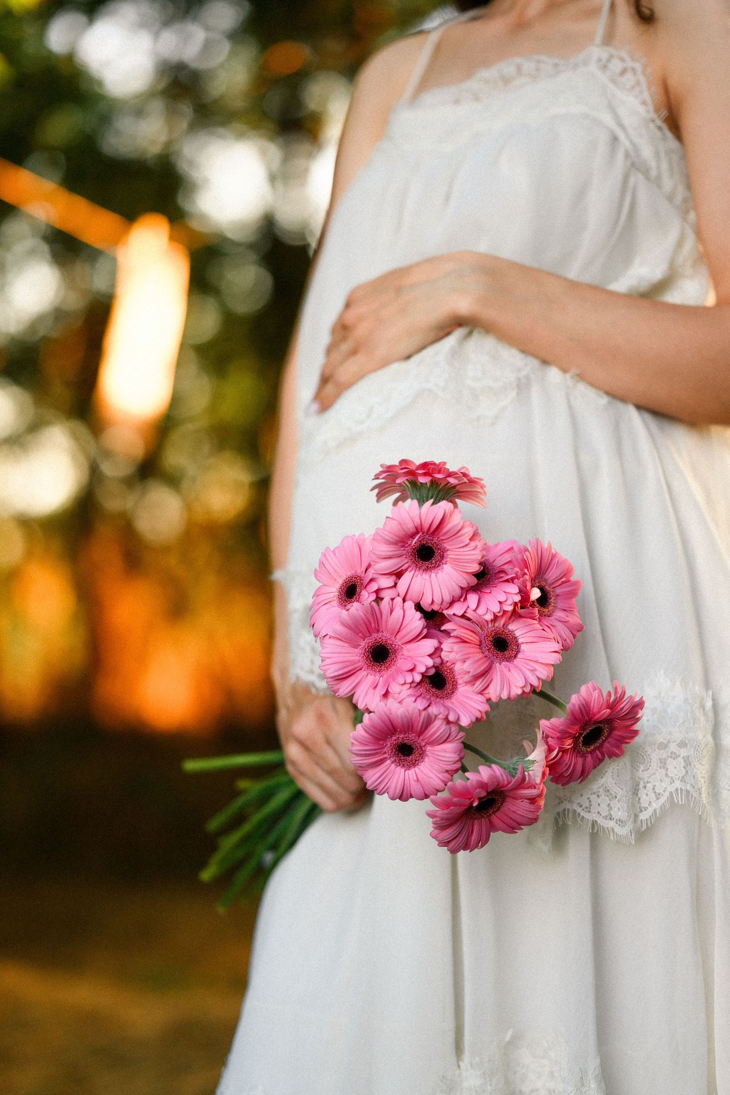 Pregnant woman in white lace dress holding a bouquet of pink gerbera daisies outdoors at golden hour.