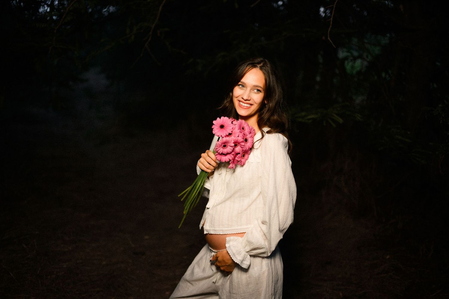 Smiling young woman in white dress holding pink flower bouquet in dark forest setting.
