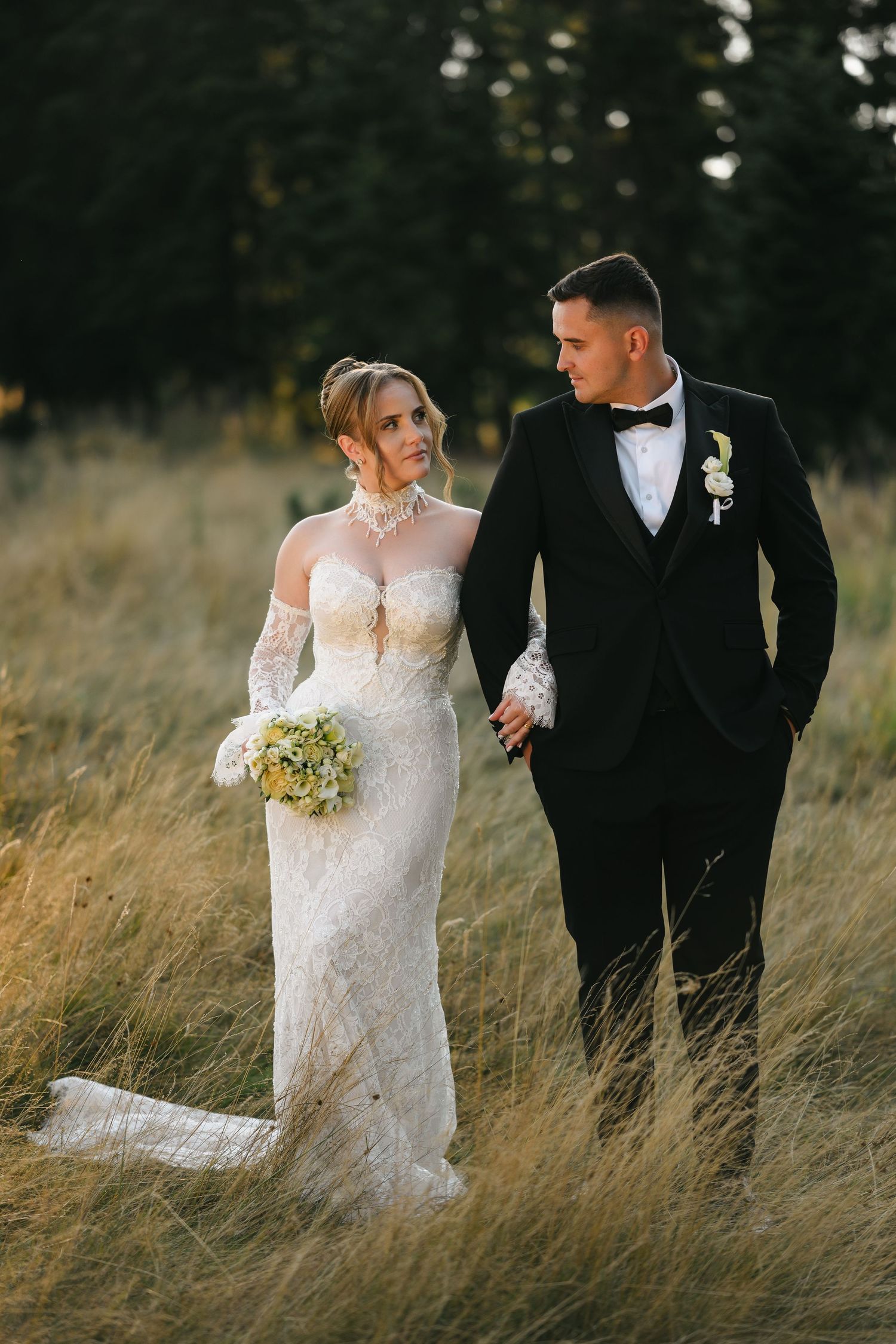 Bride in lace gown and groom in black tuxedo walk hand-in-hand through golden meadow at sunset.