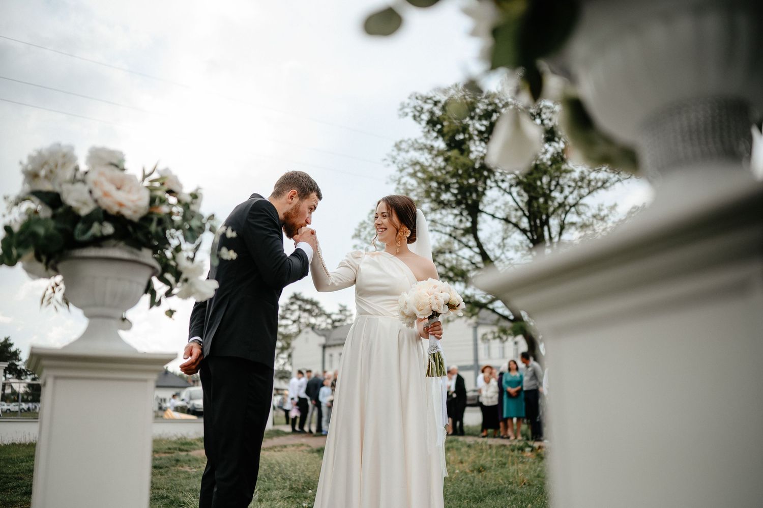 Groom tenderly kisses bride's hand during outdoor wedding ceremony with white floral decor.