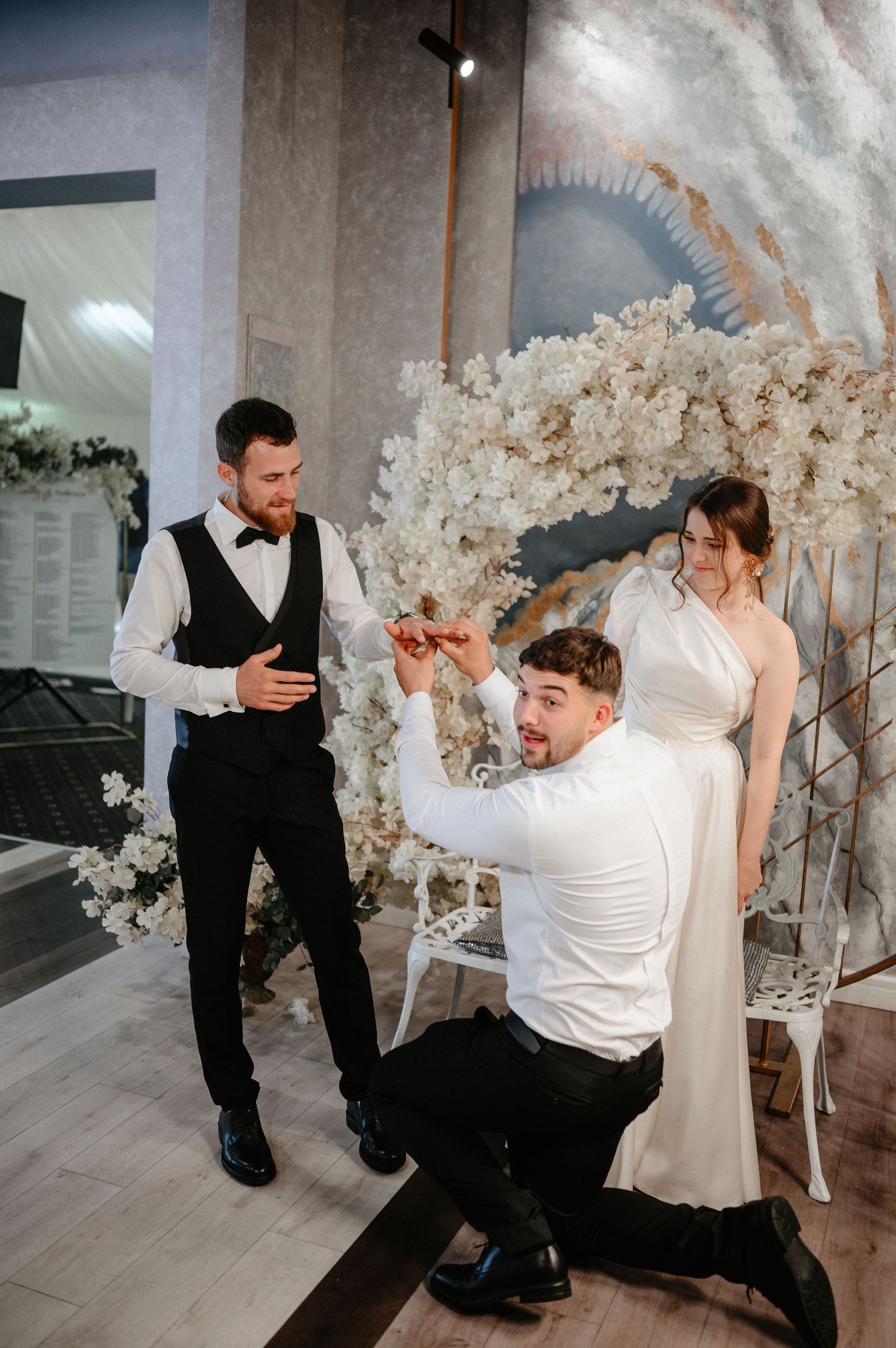 Groom kneeling to propose with ring while bride and groomsman watch at elegant white floral wedding ceremony.