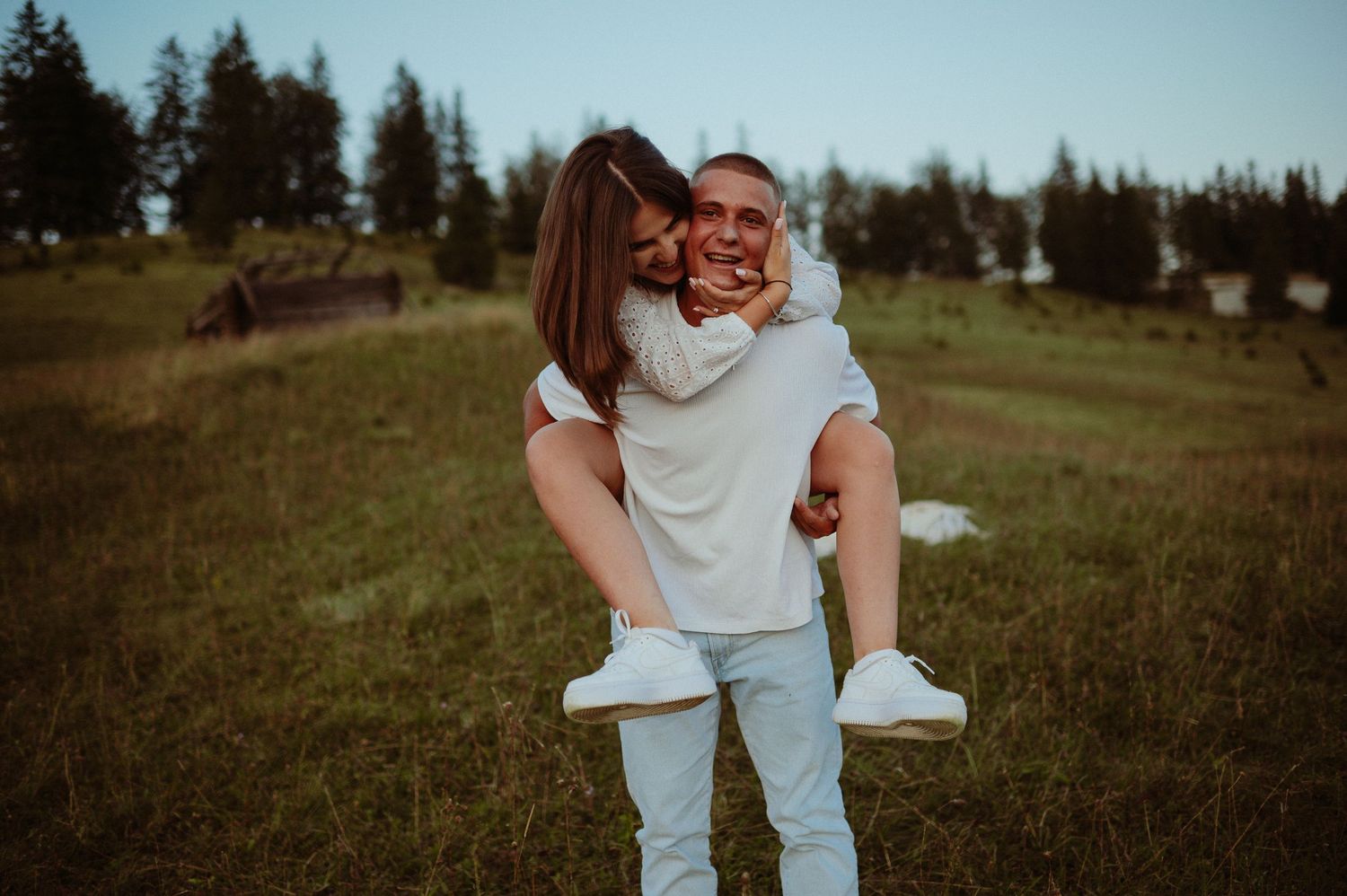 Couple sharing a kiss during piggyback ride on lush green hillside with forest backdrop at dusk.