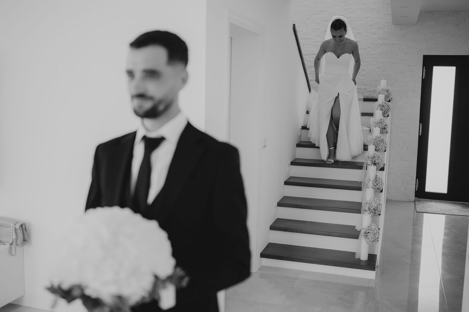 Black and white first look photo of groom holding bouquet as bride descends staircase behind him.