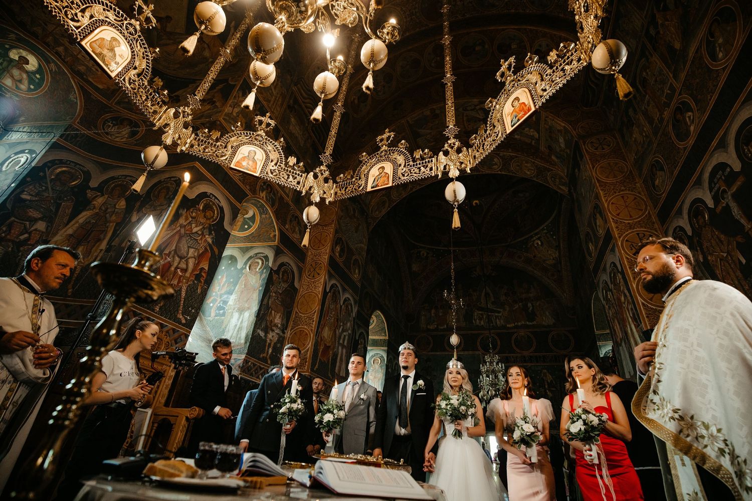 Wedding ceremony inside an ornate Orthodox church with richly decorated walls and hanging chandeliers.