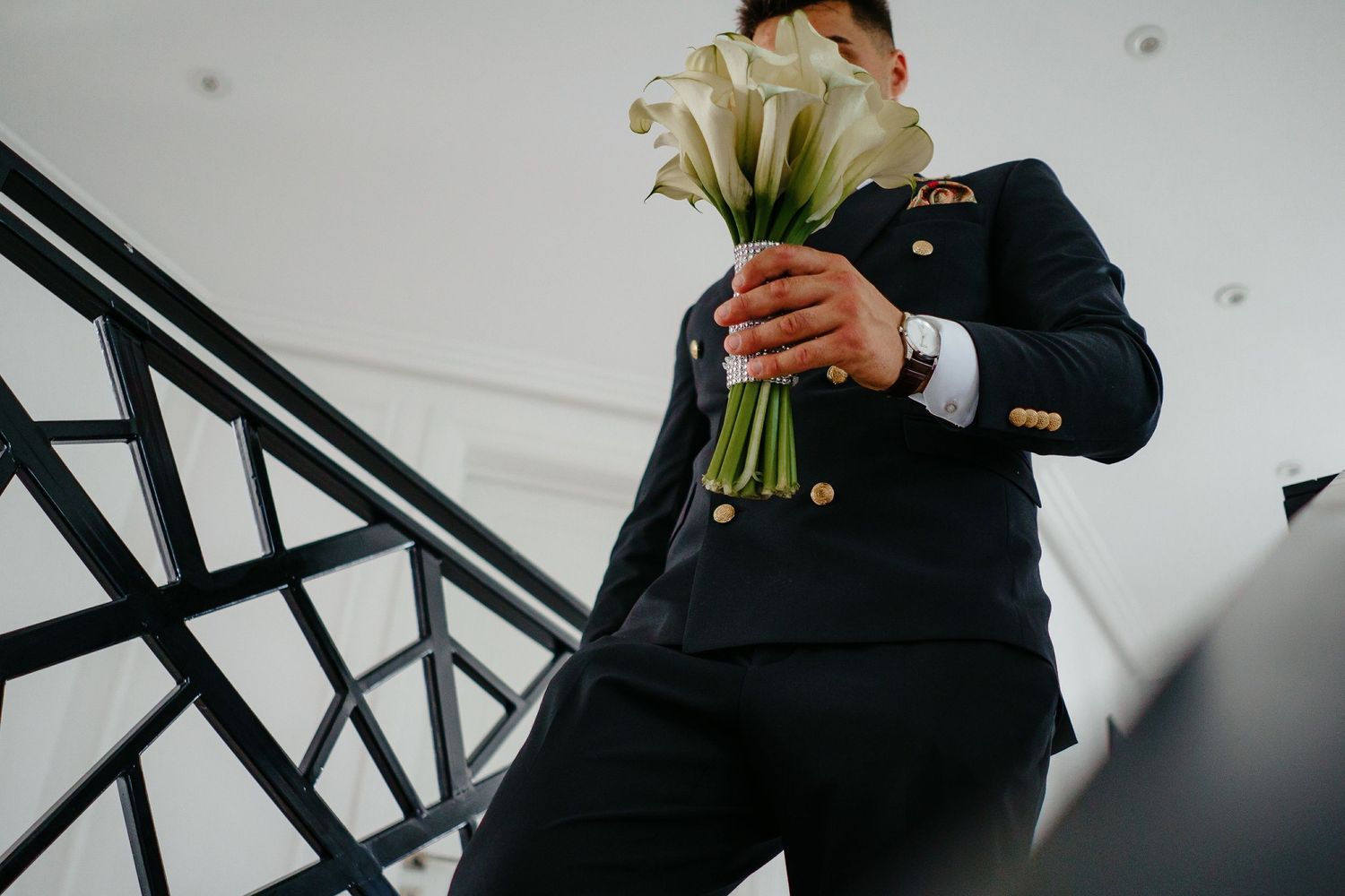 Groom holding elegant white calla lily bouquet on staircase in navy suit.