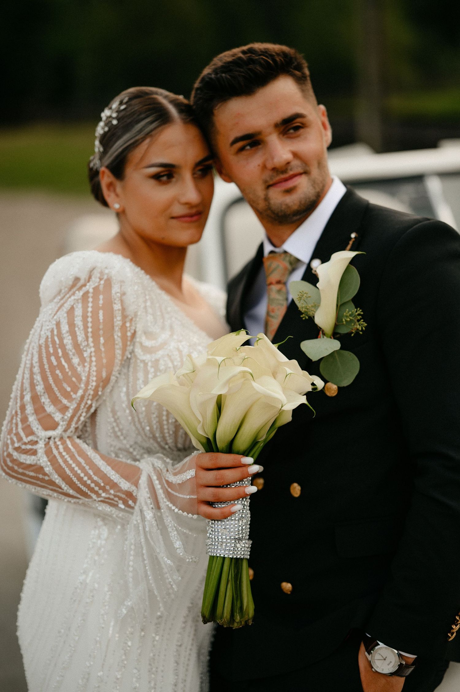 Beautiful bride holding white calla lily bouquet embraced by groom in black suit at wedding.