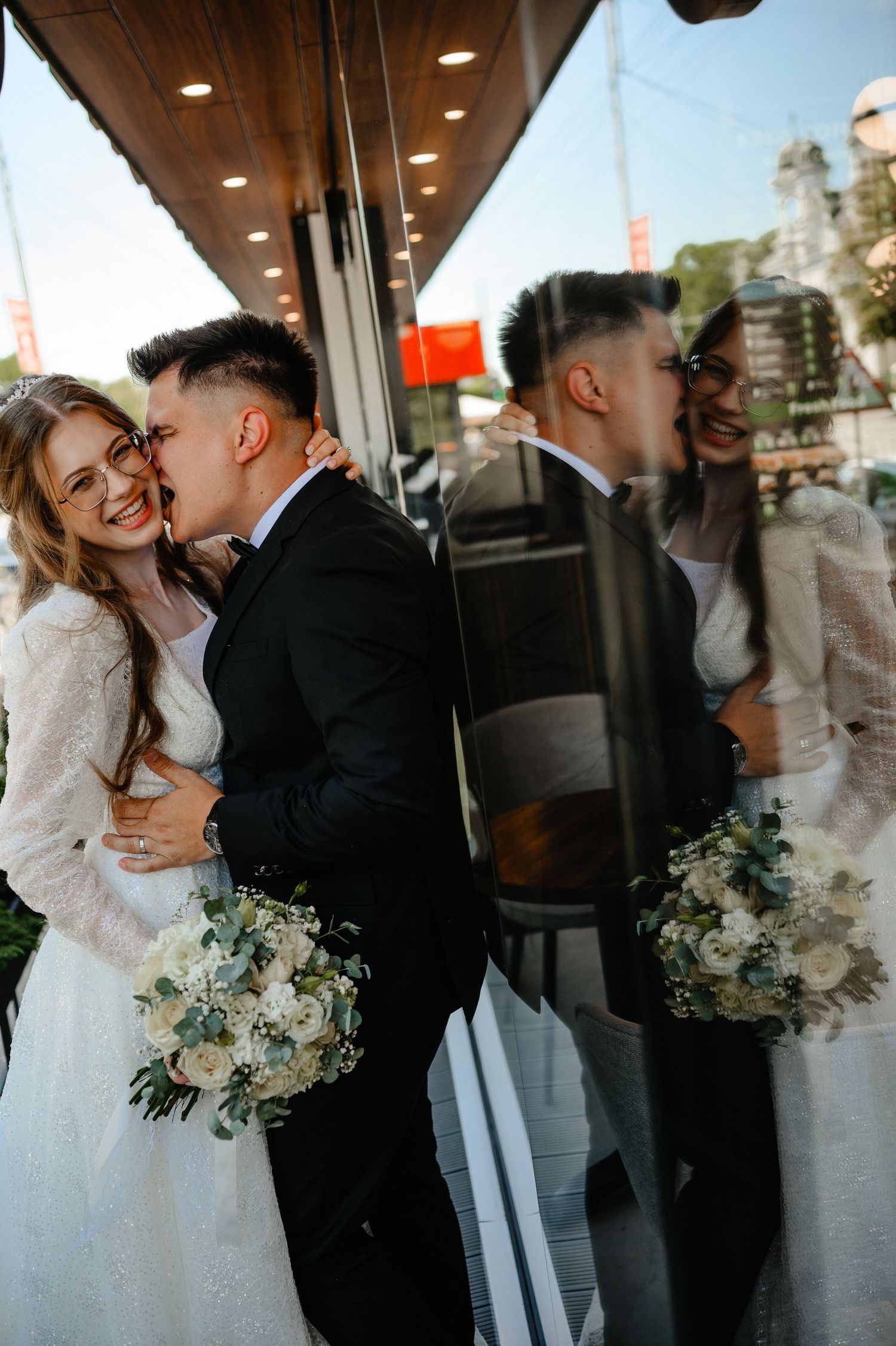 Smiling bride and groom embracing near a reflective glass window with wedding bouquet.
