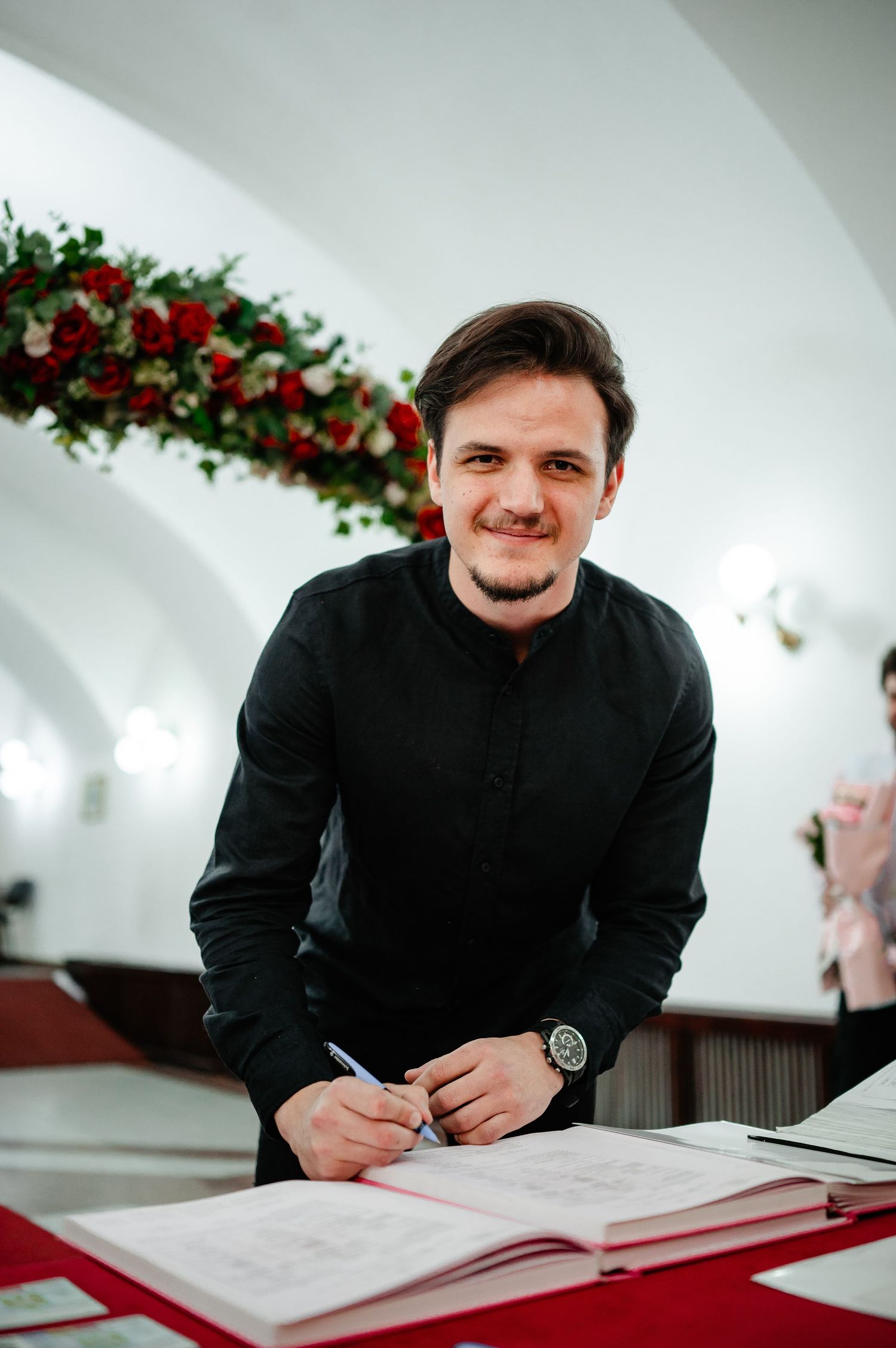 Smiling groom in black shirt signs wedding registry at red-covered table with red rose floral arch behind him.
