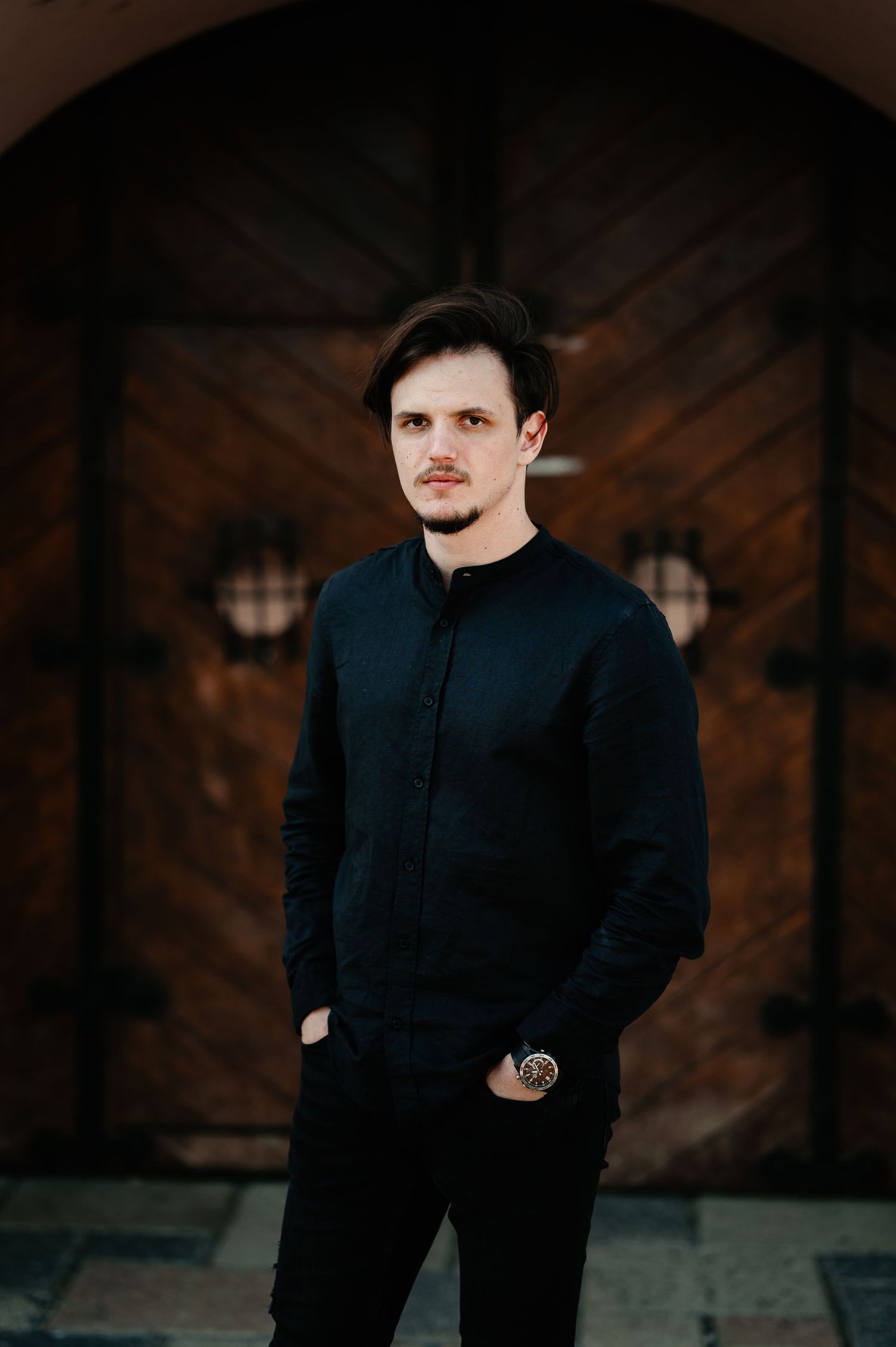 Young man in black shirt standing confidently before a rustic wooden arched door.