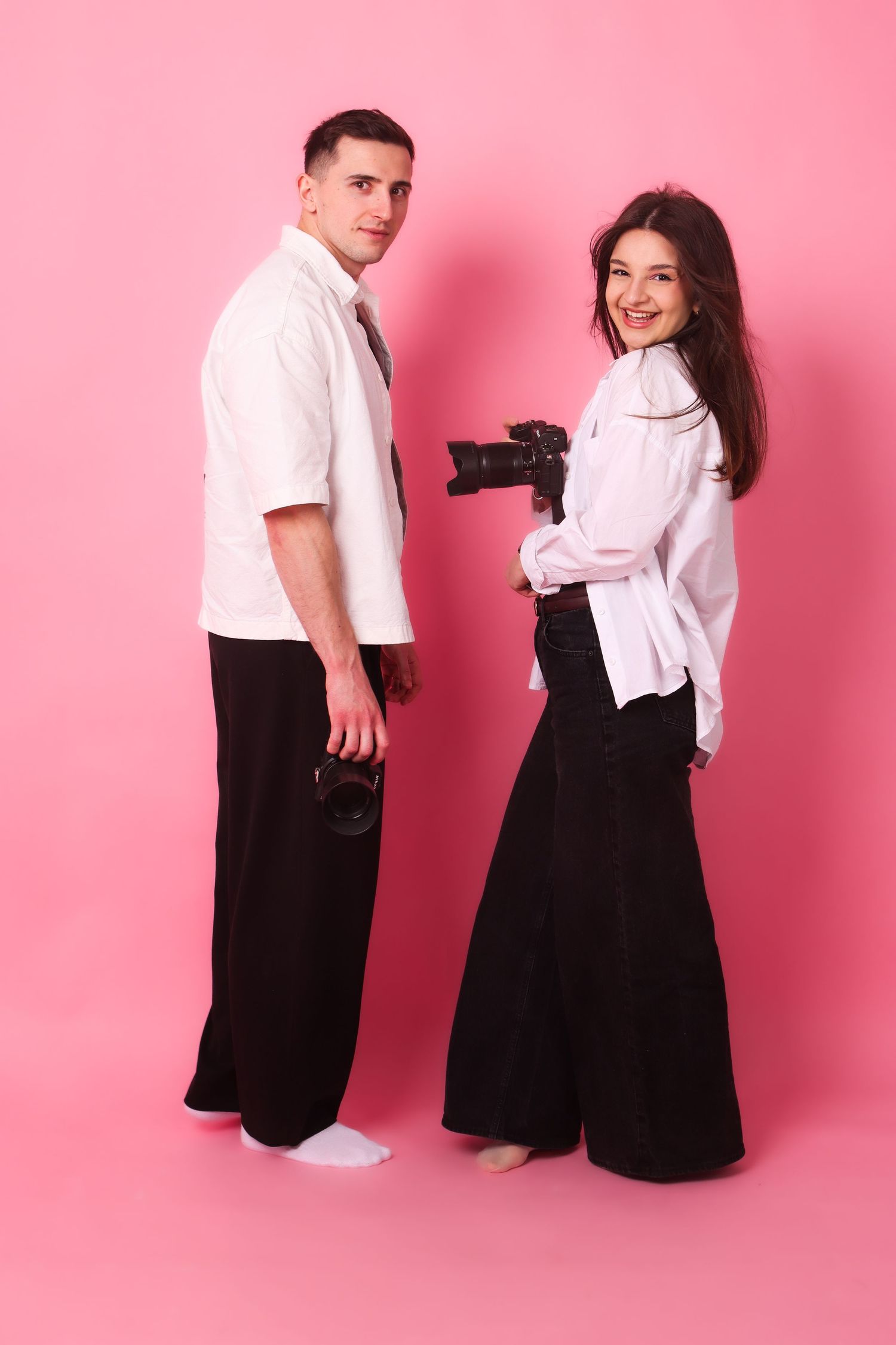 A smiling couple in matching black and white outfits hold cameras while posing on pink background.