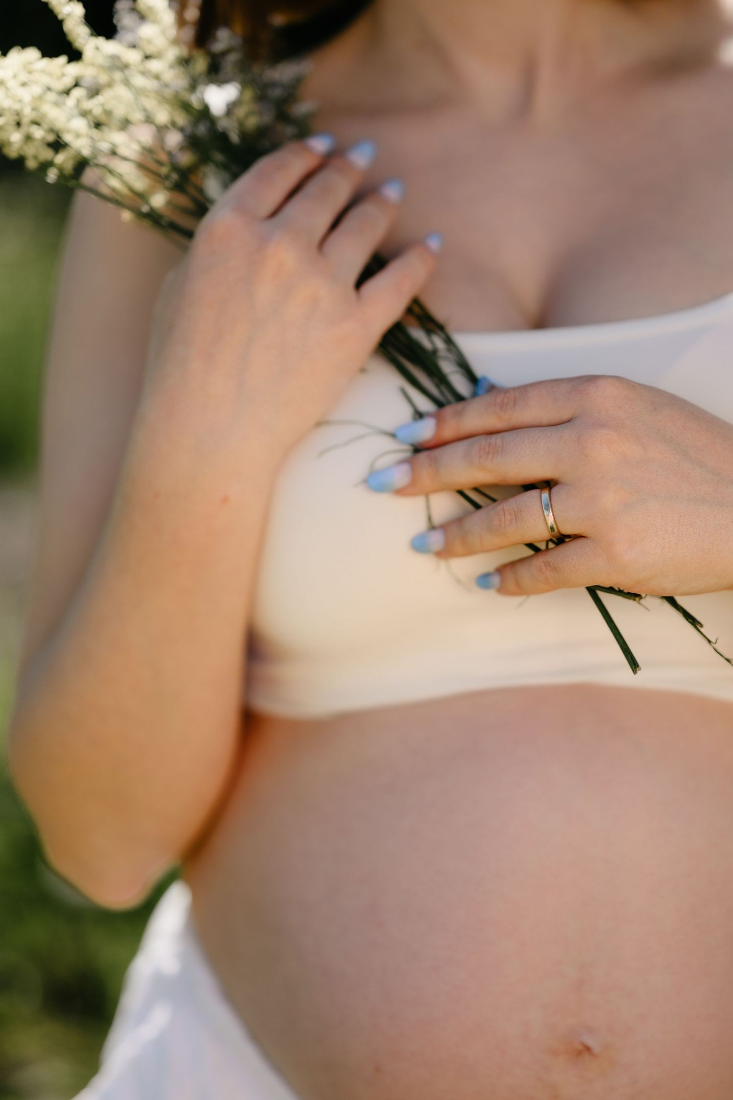 Close-up of pregnant woman in white top holding wildflowers with blue manicured nails.