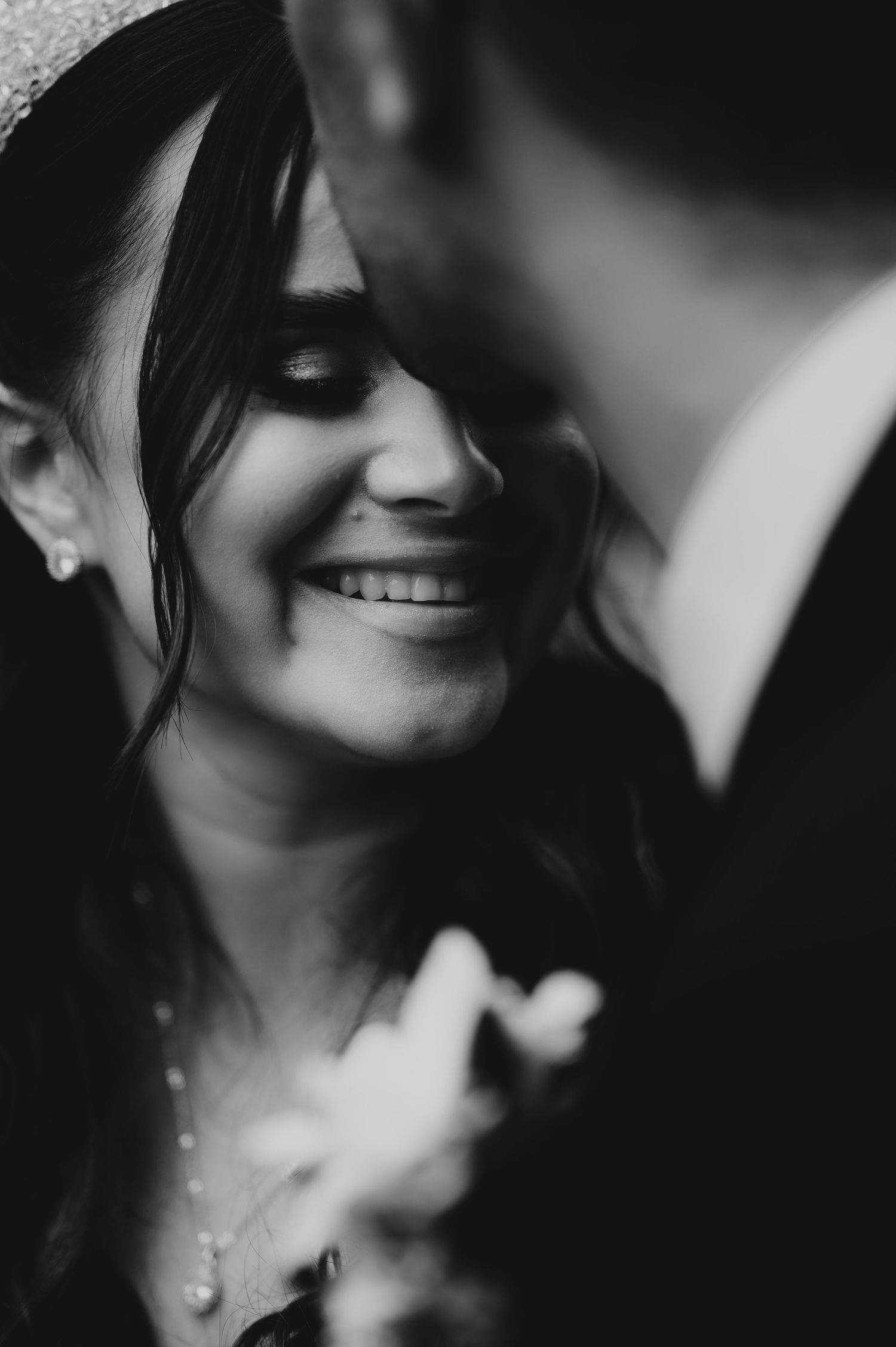 Black and white close-up of a smiling bride wearing a hat and pearl necklace holding flowers near the groom.
